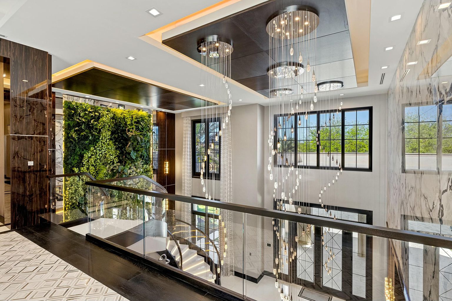 This is an interior shot of a luxurious hallway and staircase area. The space features a striking chandelier, a green wall, and a glass-enclosed staircase. The design incorporates dark wood accents, marble walls, and patterned flooring, creating a sophisticated and modern aesthetic.