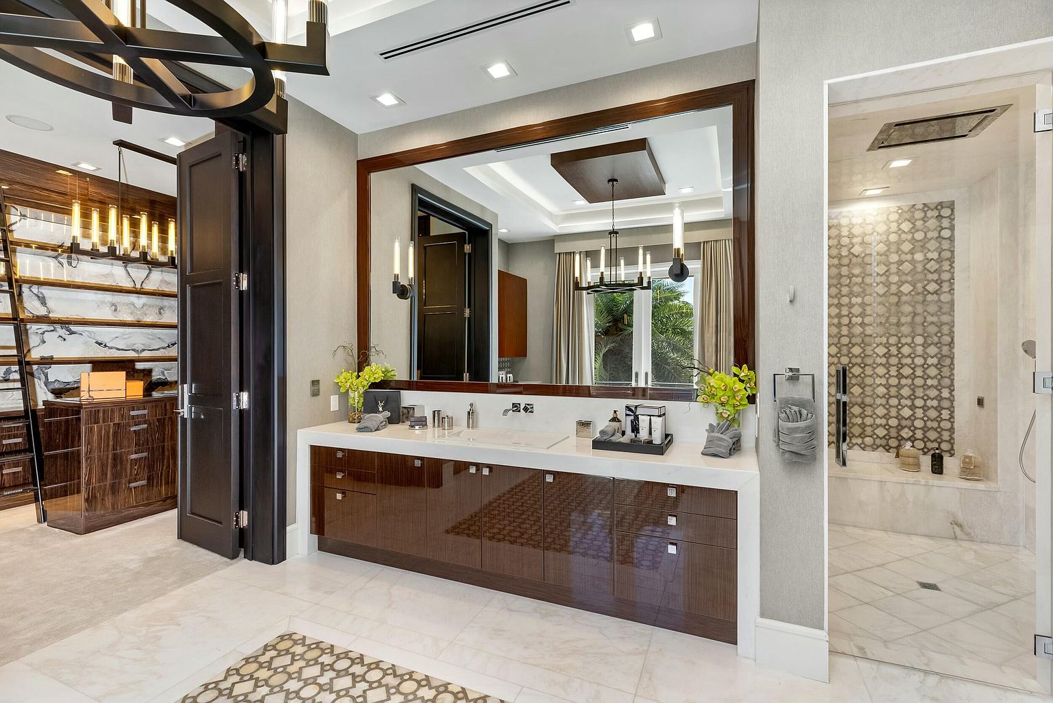 This is a luxurious primary bathroom featuring a large vanity with dark wood cabinetry and a white countertop. A large mirror reflects the room, including a glimpse of a walk-in closet and a modern shower with patterned tile. The overall impression is one of elegance and sophistication.