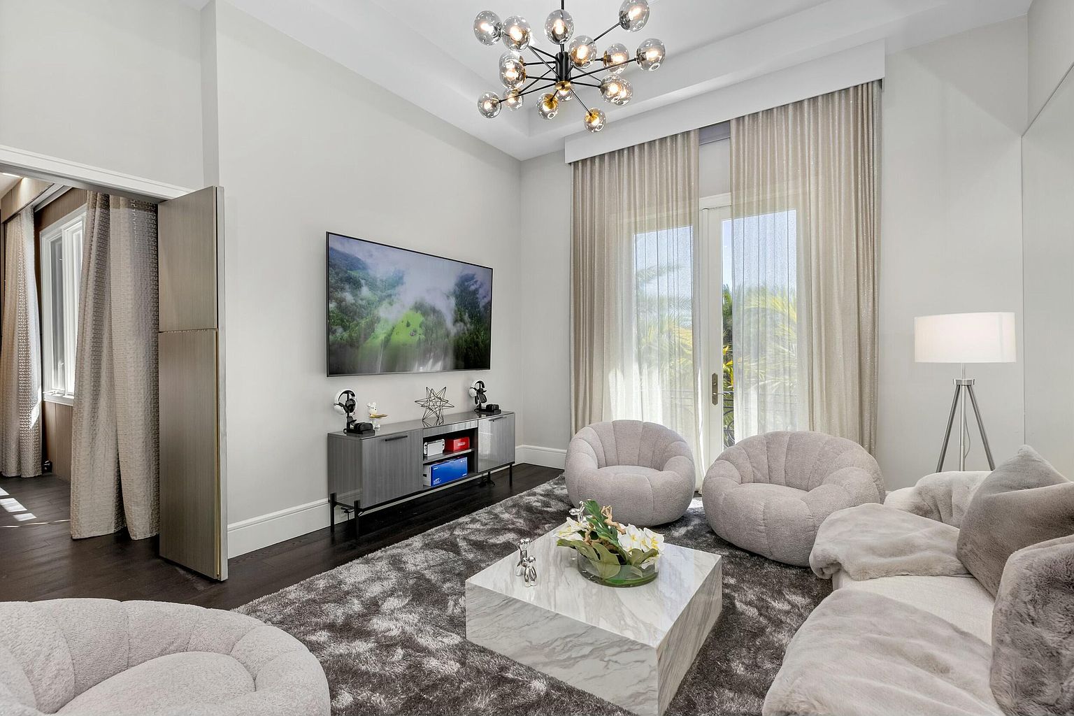 This is an interior shot of a modern living room featuring a large television mounted above a gray cabinet. The room is decorated with two round chairs, a marble coffee table with floral arrangement, and a plush gray rug. Natural light streams in through the window, complemented by a modern chandelier, creating a bright and inviting atmosphere.