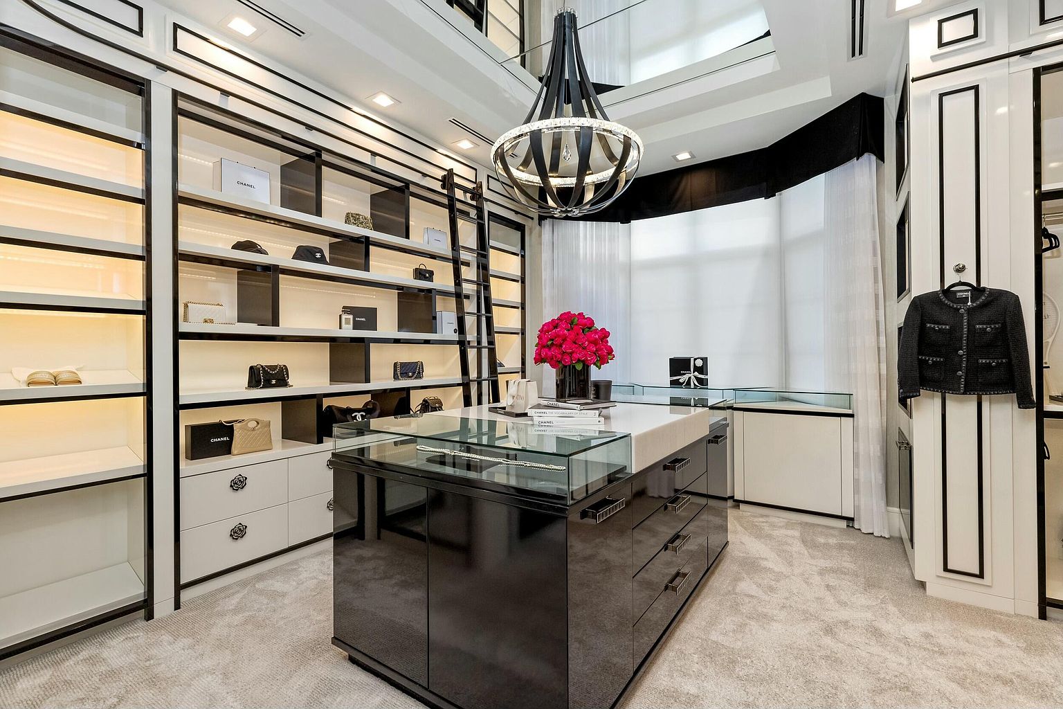 This is a luxurious walk-in closet featuring custom shelving with integrated lighting, a central island with glass display, and a rolling ladder for accessing higher shelves. The closet is decorated with designer items and a striking chandelier, creating a high-end, boutique-like atmosphere. The color palette is primarily black and white, contributing to a sophisticated and organized space.
