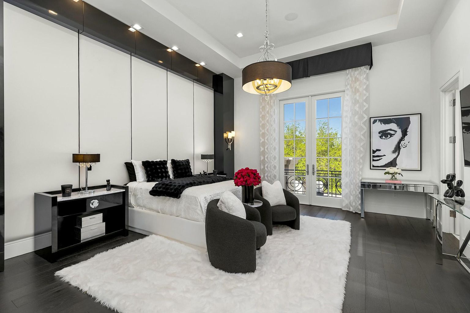 This is a luxurious primary bedroom featuring a king-size bed with black and white bedding, flanked by black nightstands with lamps. The room is decorated in a modern style with a large white shag rug, two gray armchairs, and a mirrored desk. French doors lead to a balcony, and a large portrait of Audrey Hepburn hangs on the wall.