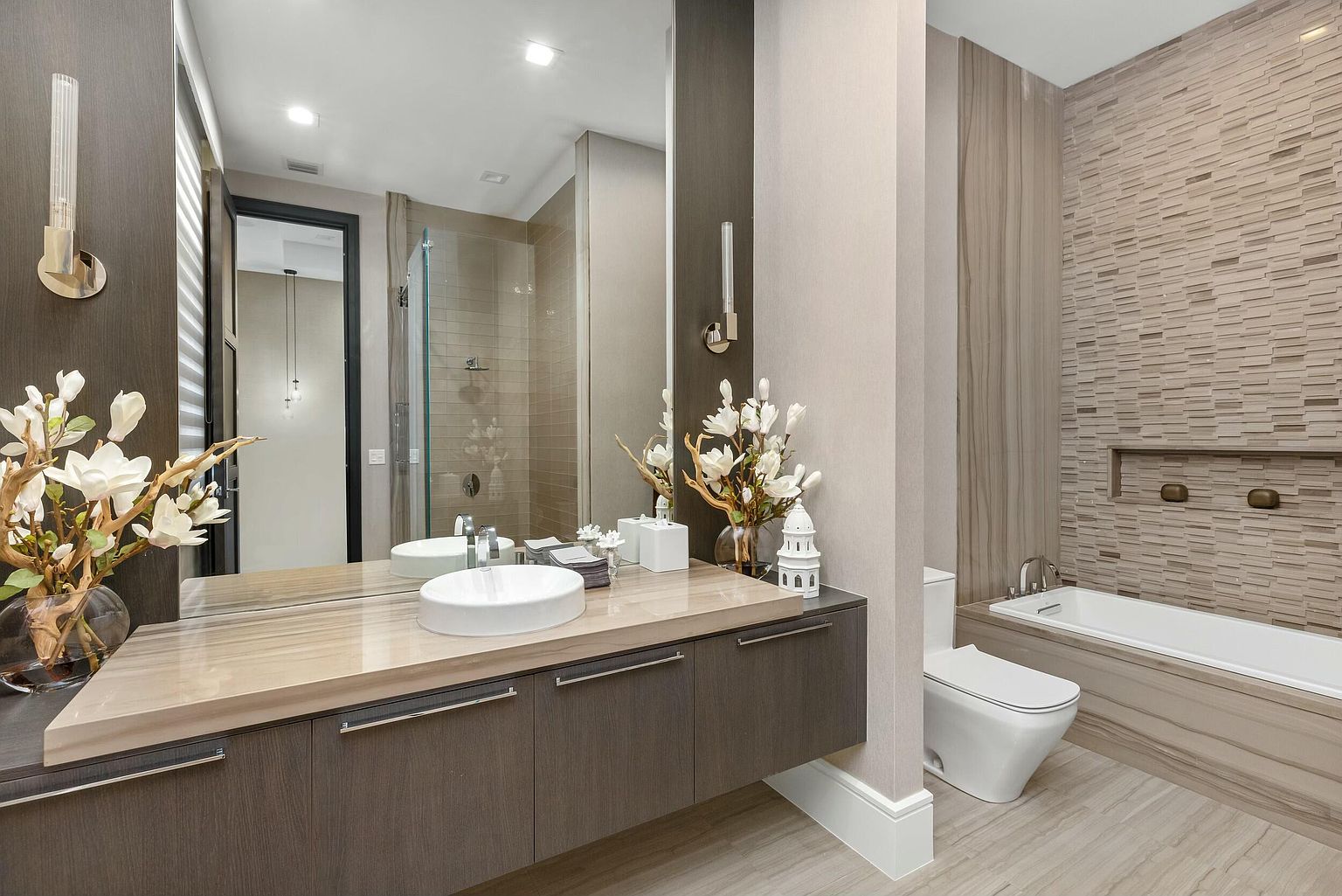 This is a modern primary bathroom featuring a floating vanity with a light-colored countertop and dark wood cabinets. A large mirror reflects the vanity and a glass-enclosed shower. The bathroom also includes a toilet and a bathtub with a tiled surround, creating a luxurious and spa-like atmosphere.