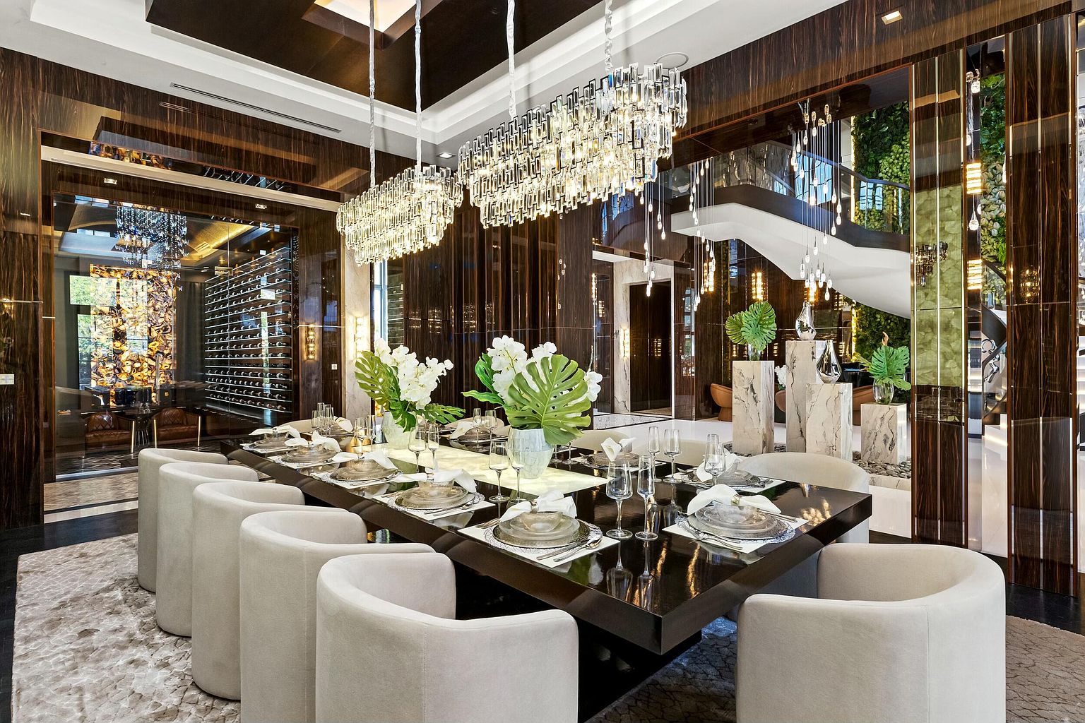 This is a luxurious dining room featuring a long, dark wood table set for a formal meal, surrounded by upholstered chairs. Above the table hang modern crystal chandeliers, and the room is decorated with elegant floral arrangements. The space is adjacent to a wine cellar and a staircase, suggesting a high-end, sophisticated home.