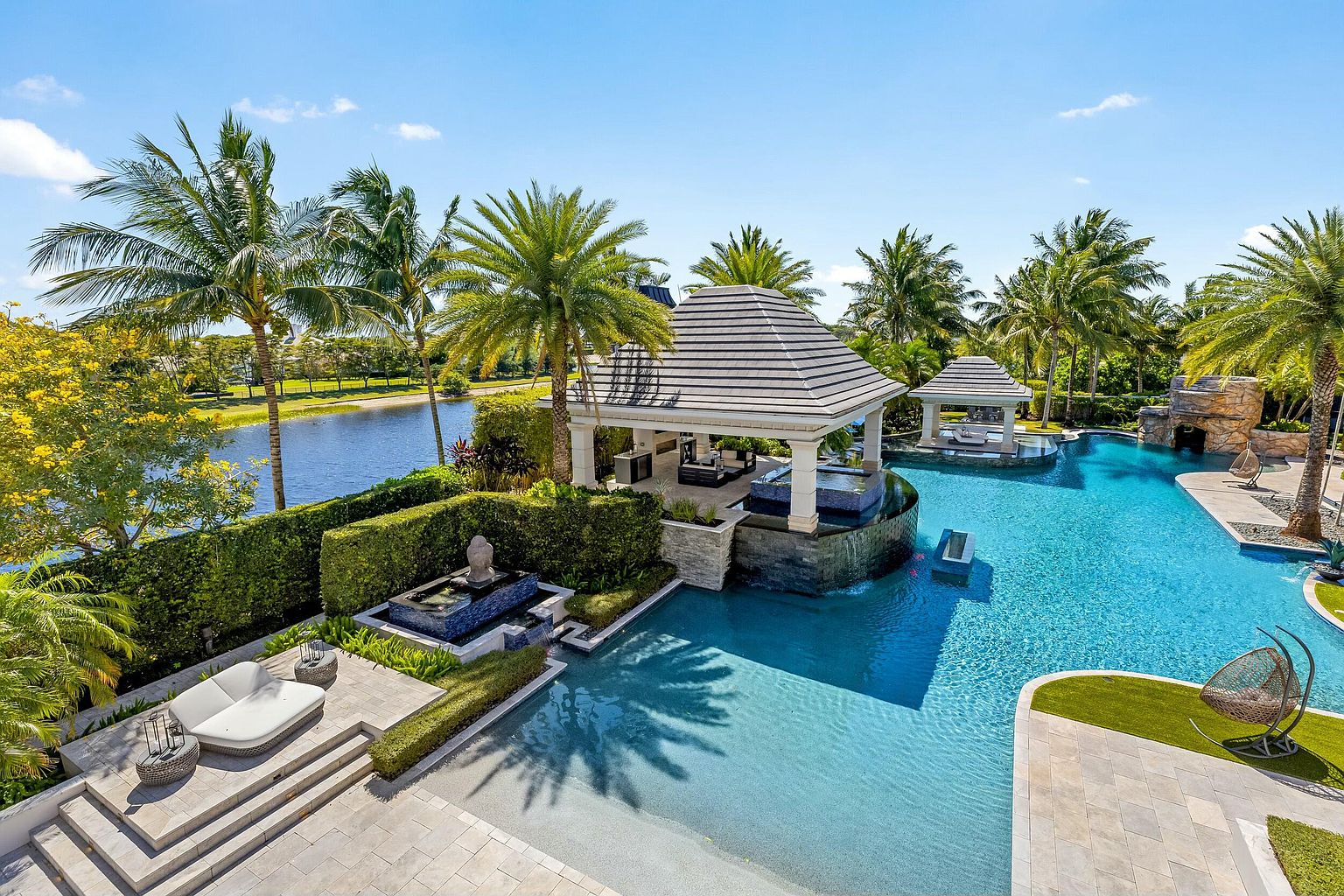 This is an aerial view of a luxurious backyard featuring a large, uniquely shaped pool with a sandy beach entry. The pool area includes two covered cabanas, one with a swim-up bar, and a rock waterfall feature. Lush tropical landscaping with palm trees surrounds the pool, creating a private and resort-like atmosphere.