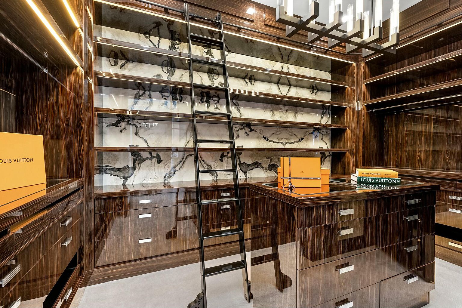 This is a luxurious walk-in closet featuring dark wood cabinetry with a high-gloss finish and marble accents. A rolling ladder provides access to the upper shelves, and the space is illuminated by modern lighting fixtures. The closet exudes sophistication and offers ample storage.