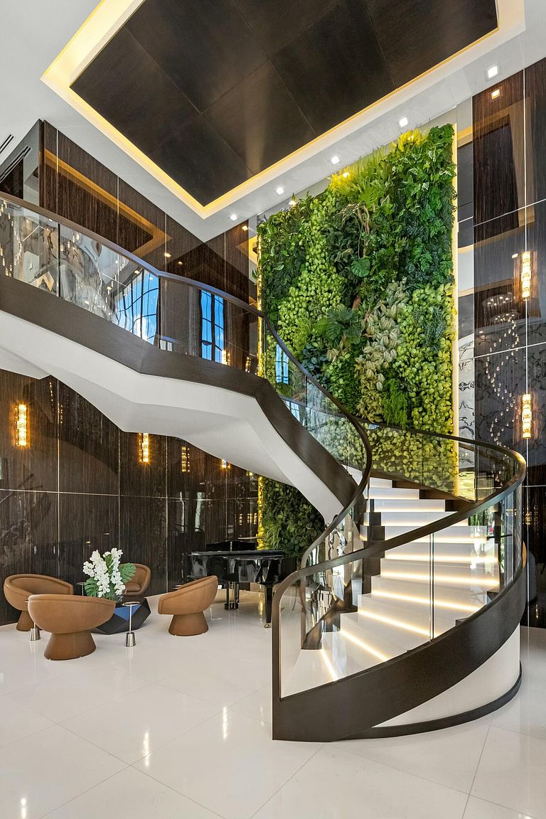 This grand interior showcases a stunning spiral staircase with illuminated steps and a glass railing, complemented by a striking vertical garden wall. The space features dark, glossy wall panels, a black grand piano, and modern seating, creating a luxurious and sophisticated ambiance. The high ceiling with unique lighting fixtures adds to the overall elegance.