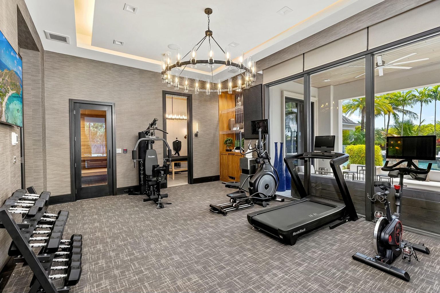 This is a well-equipped home gym featuring a variety of exercise machines including a treadmill, elliptical, stationary bike, and a multi-station weight machine. A rack of dumbbells is also present. The room has a neutral color palette with textured wallpaper and a large chandelier, creating a sophisticated workout environment with views to the outside.