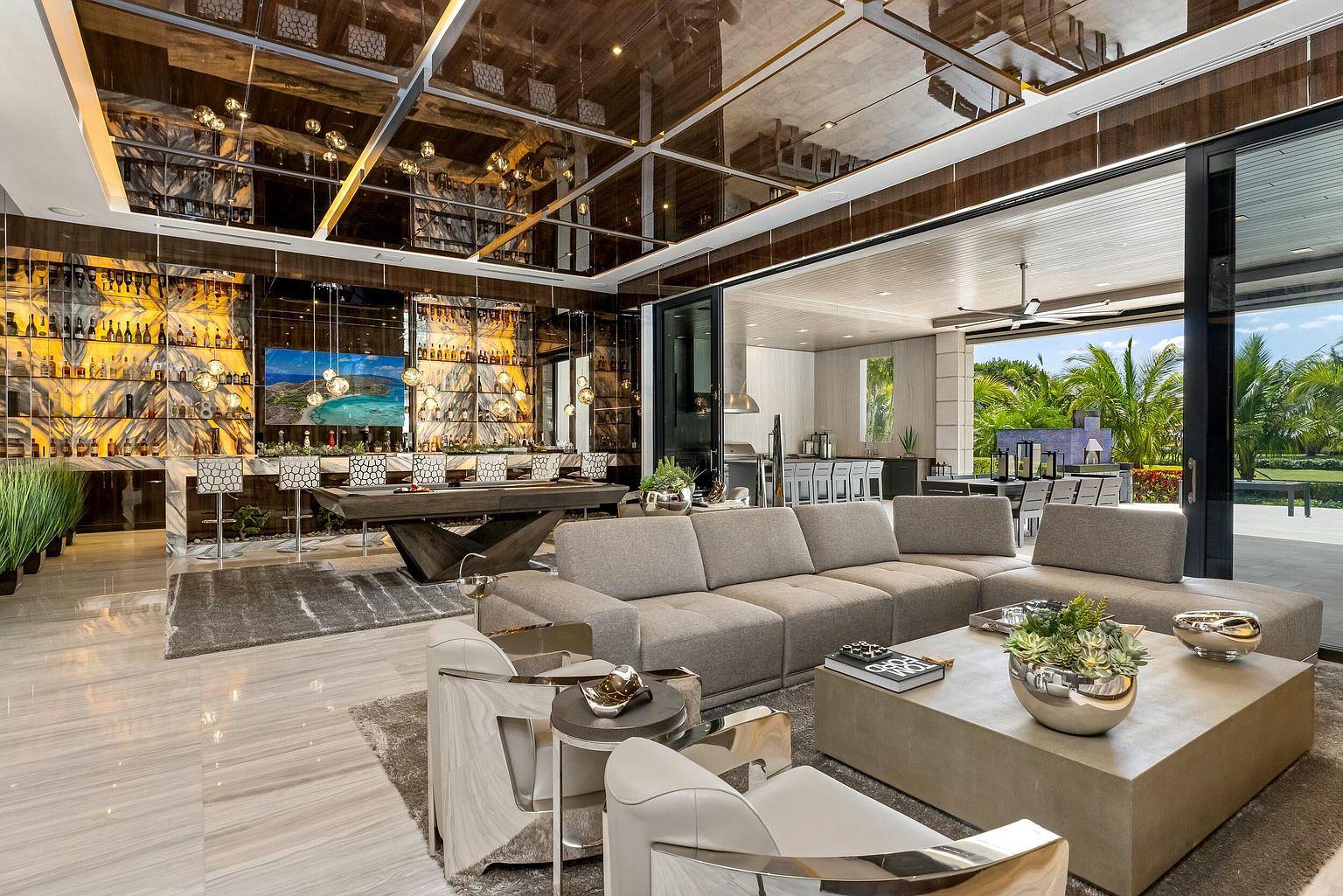 This is a luxurious living room featuring a full bar with an extensive liquor collection, a pool table, and a large sectional sofa. The room opens to an outdoor area through sliding glass doors, offering a seamless indoor-outdoor living experience. The mirrored ceiling and marble floors add to the opulent feel of the space.