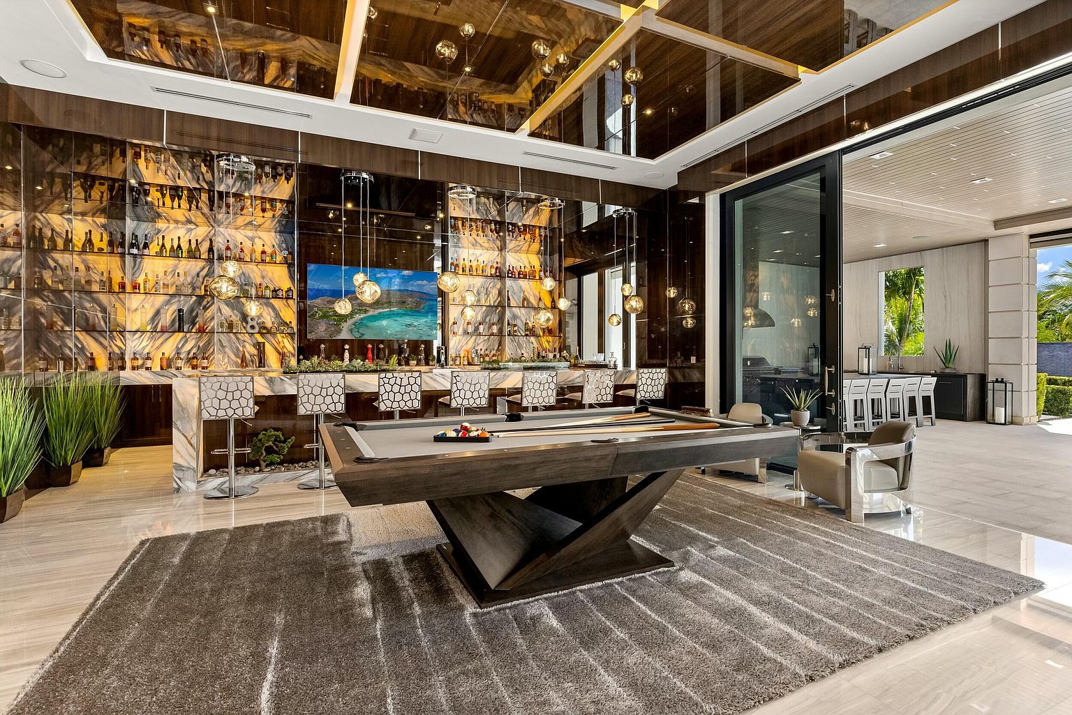 This interior shot showcases a luxurious living room featuring a modern pool table on a large rug, a fully stocked bar with stylish seating, and a mirrored ceiling with pendant lighting. The open floor plan leads to an outdoor area, creating a seamless indoor-outdoor living experience. The room exudes sophistication and entertainment.