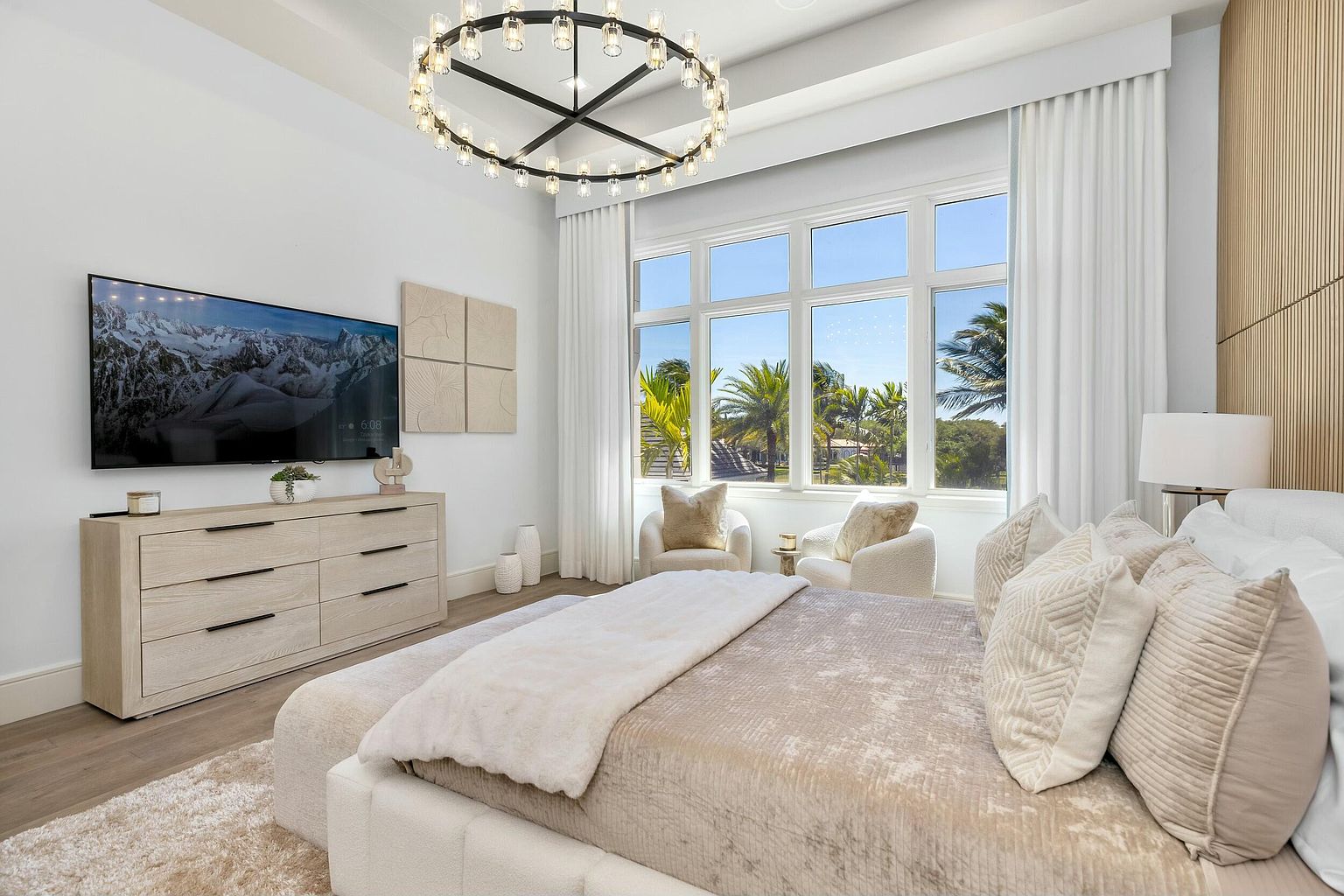This is a bright and luxurious primary bedroom featuring a large window with a view of palm trees, a modern chandelier, and a neutral color palette. The room includes a light wood dresser with a television above, two armchairs by the window, and a plush bed with decorative pillows. The overall impression is one of comfort and sophistication.