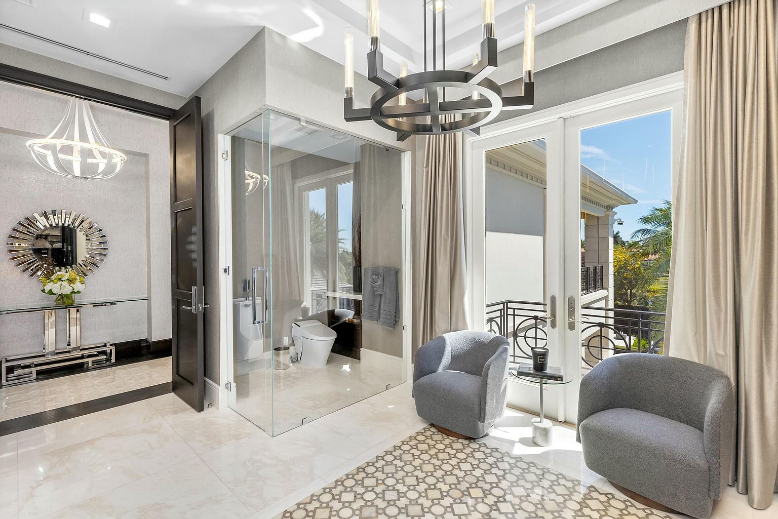 This is a luxurious primary bathroom featuring a glass-enclosed shower, a modern chandelier, and two comfortable gray armchairs. The room has a neutral color palette with marble flooring and elegant fixtures. Large windows provide natural light and a view to the outside, enhancing the spa-like atmosphere.