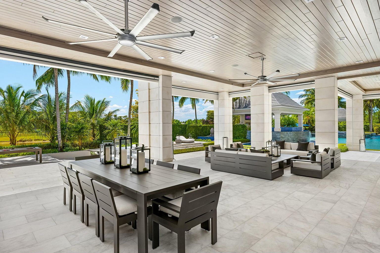 This image showcases a luxurious outdoor patio area, featuring a large dining table with seating, a comfortable lounge area with sofas and chairs, and elegant architectural columns. The space is well-lit with ceiling fans and offers a view of a beautifully landscaped yard with palm trees and a pool. The overall impression is one of sophisticated outdoor living and relaxation.
