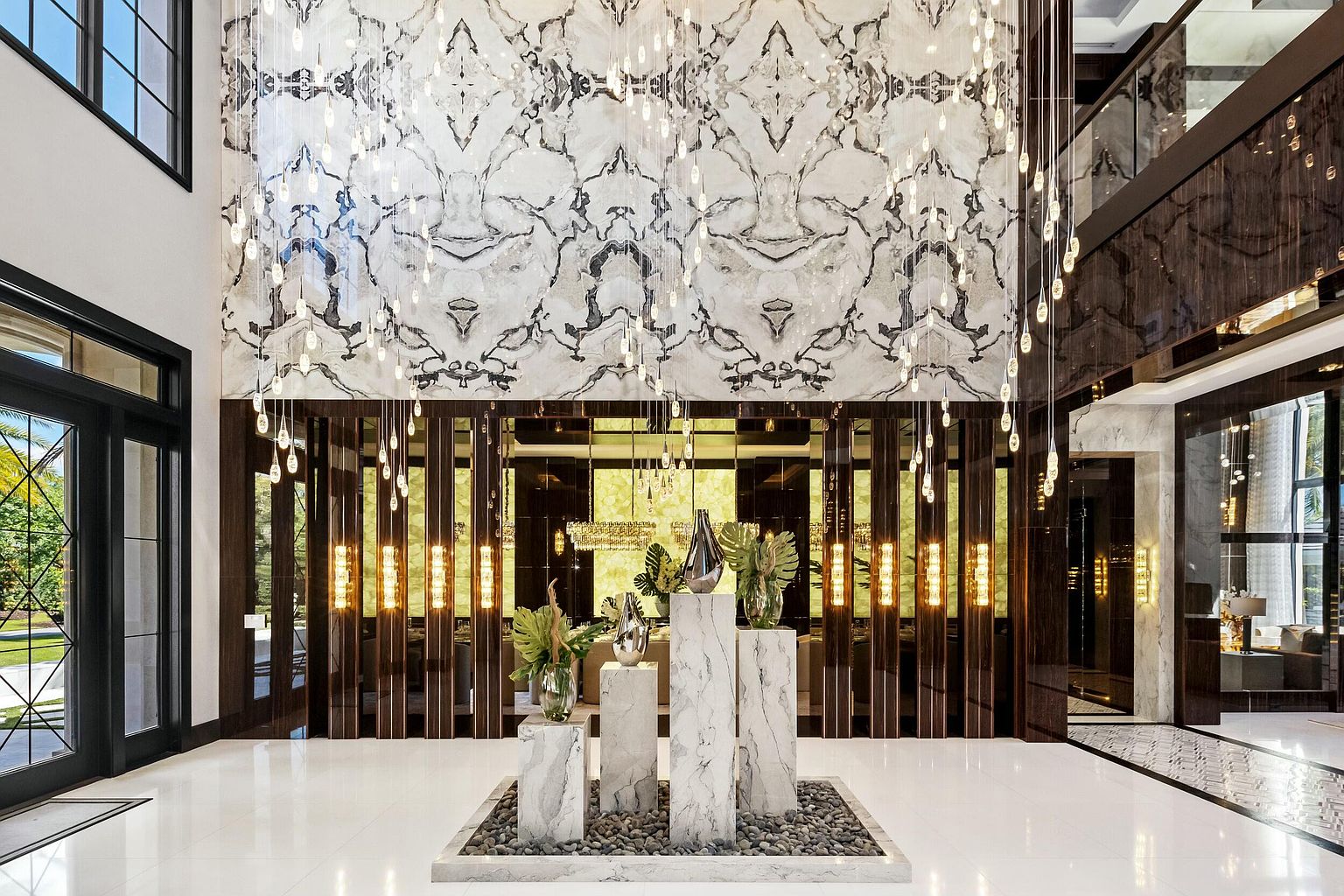 This grand foyer features a striking marble accent wall and a dazzling chandelier that creates a luxurious ambiance. The polished white floors reflect the light, enhancing the spacious feel, while the dark wood accents add a touch of sophistication. The overall impression is one of opulence and modern elegance, perfect for a high-end real estate listing.