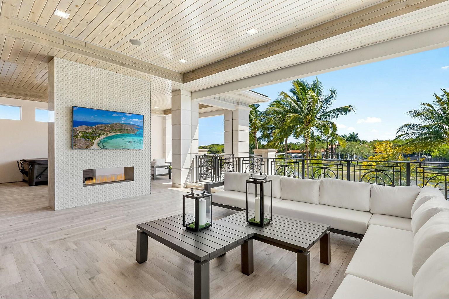 This image showcases a luxurious outdoor living space, featuring a spacious patio or balcony with elegant white sectional seating and dark wood coffee tables adorned with decorative lanterns. The area is partially covered, with a modern fireplace and TV mounted on a textured wall, offering both comfort and entertainment. The view extends to lush greenery and palm trees, creating a serene and upscale atmosphere.