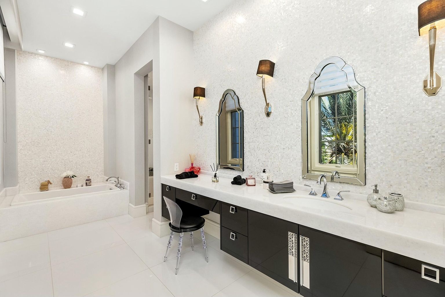 This is a luxurious primary bathroom featuring a long vanity with a white countertop and dark cabinetry. The walls are adorned with a textured, mosaic-like tile, and decorative sconces flank the mirrors. A soaking tub is visible in the background, contributing to the spa-like atmosphere of the space.