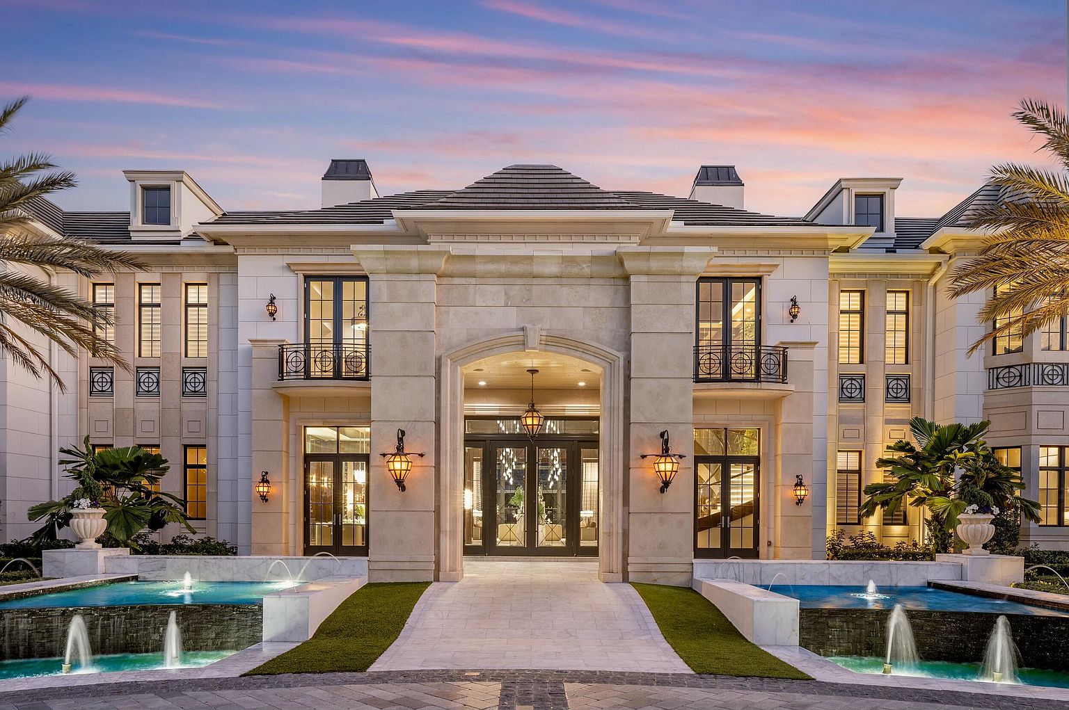 This is a grand front view of a luxurious mansion at dusk, featuring a symmetrical facade with a prominent stone entryway and double doors. The property boasts manicured landscaping, including water features with fountains, and elegant exterior lighting that enhances its curb appeal. The overall impression is one of opulence and sophisticated design.