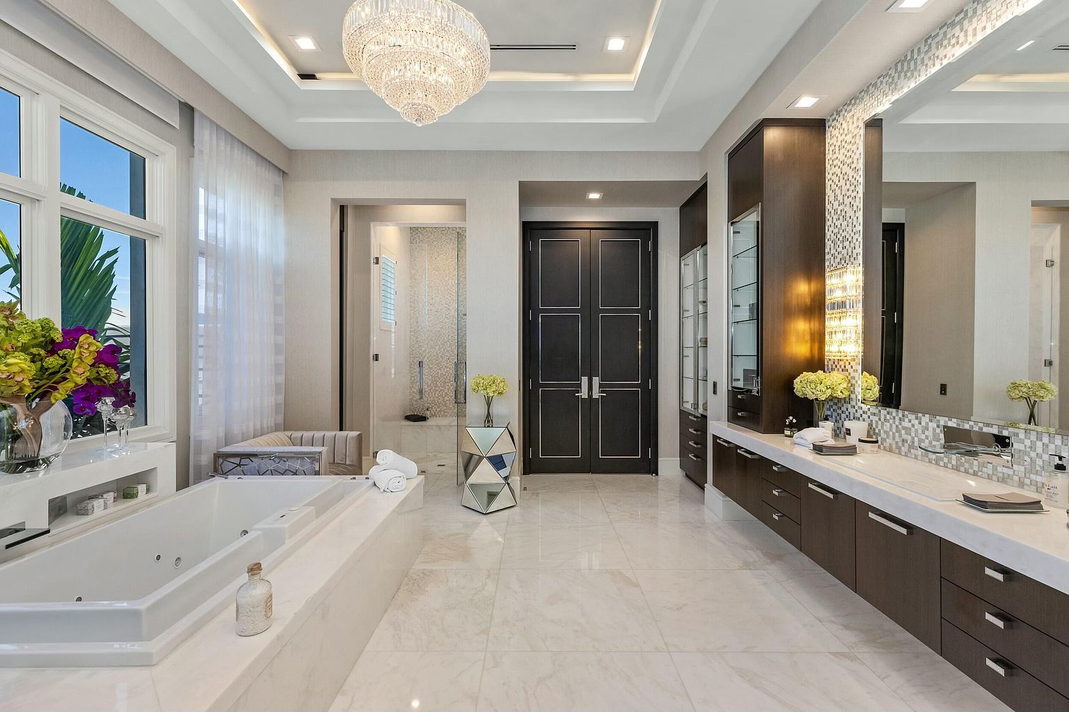 This is a luxurious primary bathroom featuring a large soaking tub, a double vanity with dark wood cabinetry and white countertops, and a separate shower area. The room is illuminated by a crystal chandelier and recessed lighting, creating a bright and elegant atmosphere. The flooring is light-colored tile, and there are decorative accents such as floral arrangements and mosaic tile details.
