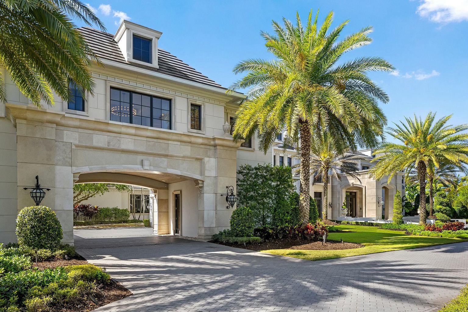 This is a front exterior view of a luxurious estate, showcasing a grand entrance with a porte-cochère. The architecture features light-colored stone, elegant windows, and meticulously landscaped grounds with mature palm trees. The driveway is paved with interlocking stones, leading to a covered entrance, creating an inviting and impressive approach to the property.