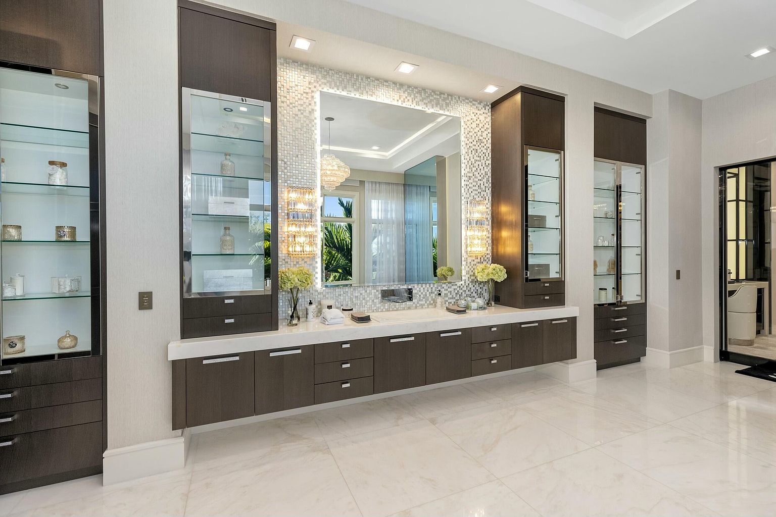 This is a luxurious primary bathroom featuring a long vanity with dark wood cabinetry and a white countertop. A large, mosaic-tiled mirror is centered above the vanity, flanked by glass-fronted display cabinets. The room is brightly lit with recessed lighting and decorative sconces, and the floor is covered in large, light-colored tiles, creating a clean and spacious feel.