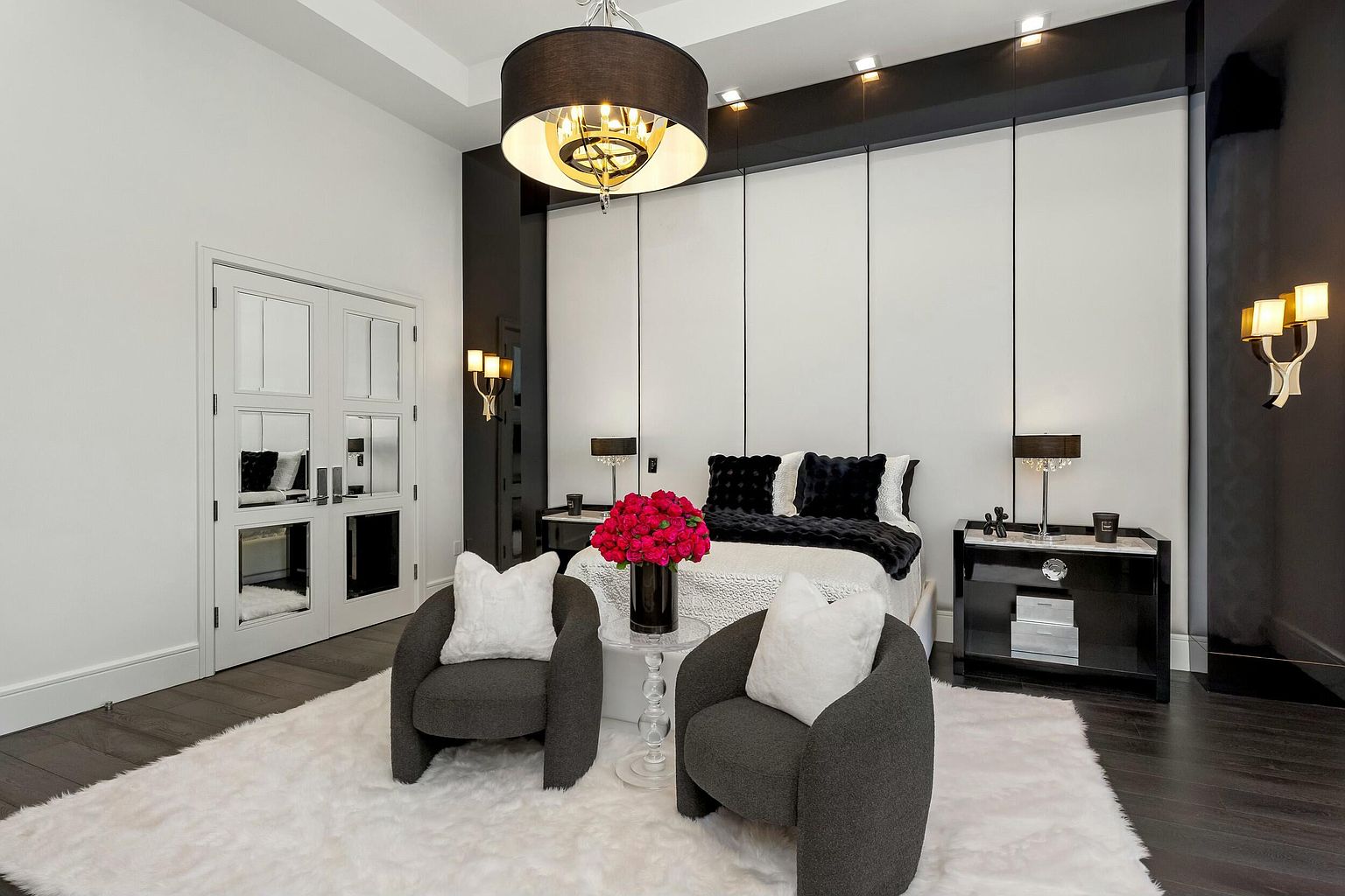 This is a luxurious primary bedroom featuring a king-size bed with black and white bedding, flanked by matching black nightstands and lamps. Two gray armchairs with white pillows sit on a plush white rug in the foreground, adding a cozy seating area. The room is decorated in a modern style with black and white accents, including a striking black and white paneled headboard and a statement chandelier.