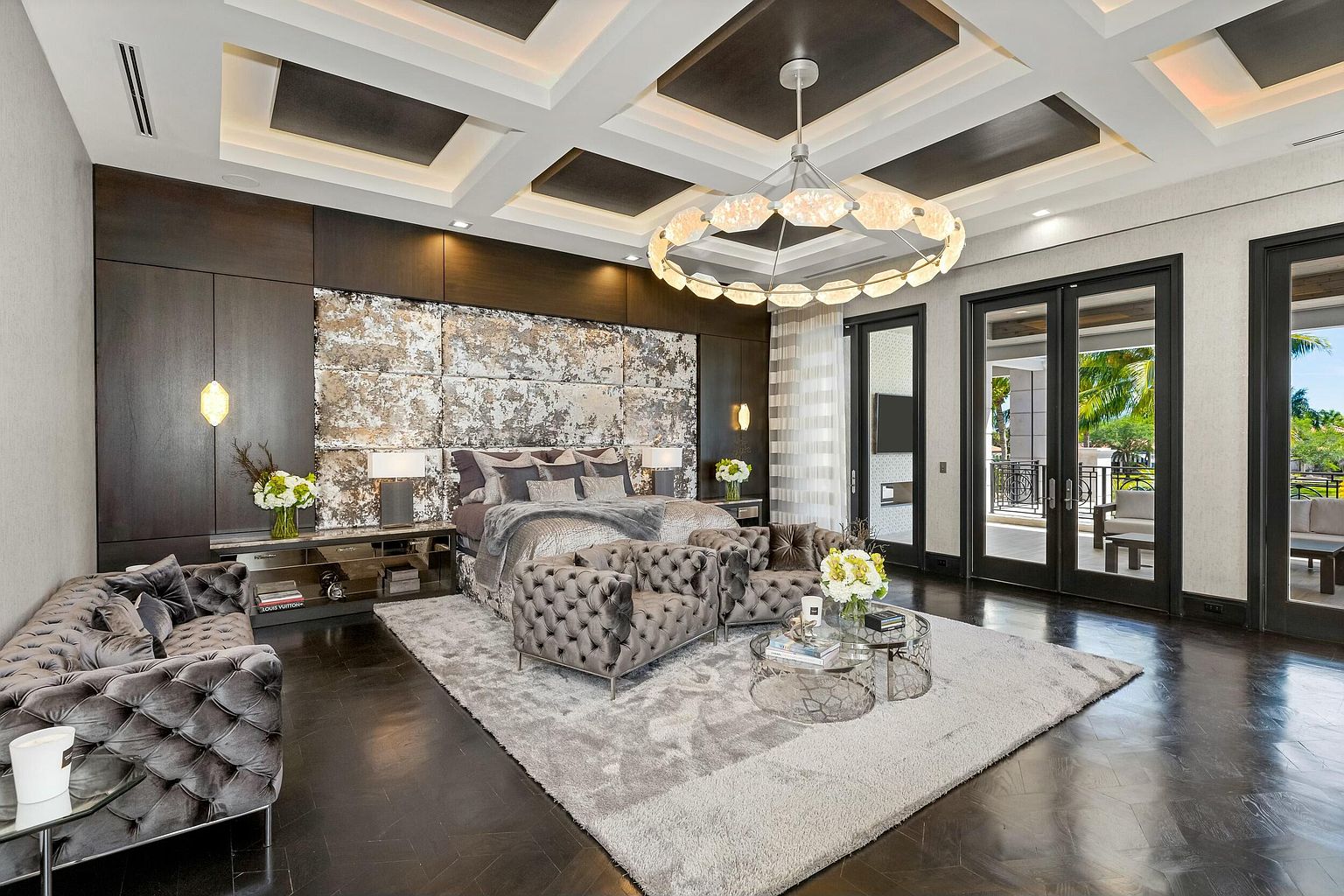 This is a luxurious primary bedroom featuring a large bed with a textured headboard, flanked by mirrored nightstands. The room is decorated in neutral tones with gray tufted seating and a large area rug. A modern chandelier hangs from the coffered ceiling, and sliding glass doors lead to an outdoor balcony.