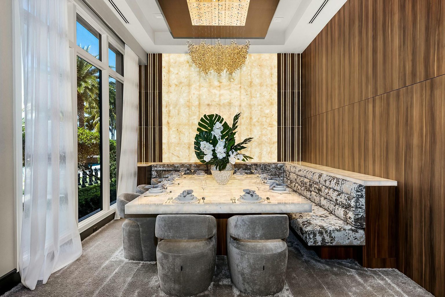This is an interior shot of a luxurious dining room. The room features a large marble dining table set for a meal, surrounded by unique gray chairs and a built-in upholstered bench. A stunning chandelier hangs above the table, and a large window provides natural light, creating an elegant and inviting atmosphere.