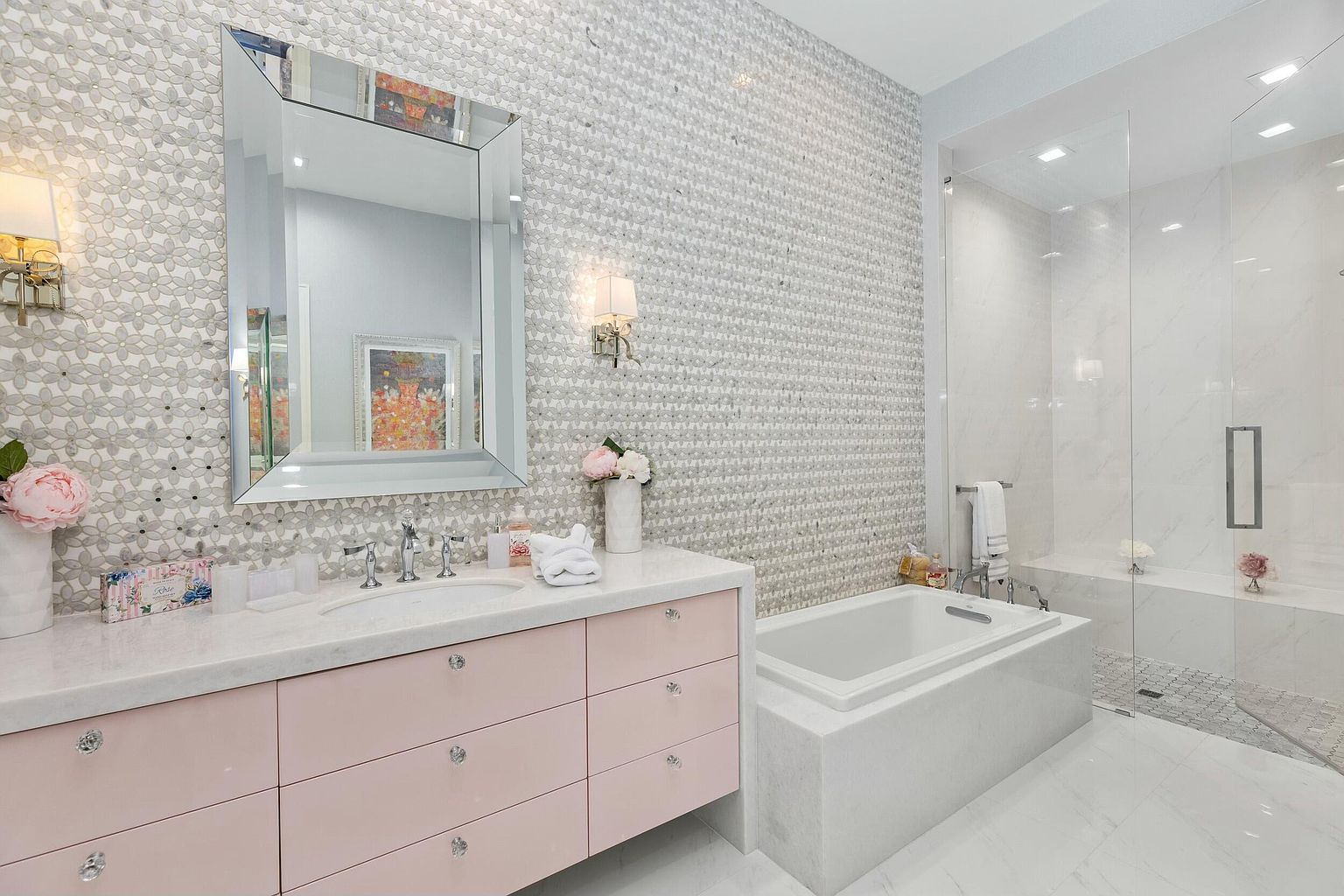 This is a luxurious primary bathroom featuring a pink vanity with crystal knobs, a marble countertop, and a large, framed mirror. The walls are adorned with intricate mosaic tiles, and a freestanding tub is situated next to a glass-enclosed shower. The overall impression is elegant and sophisticated.