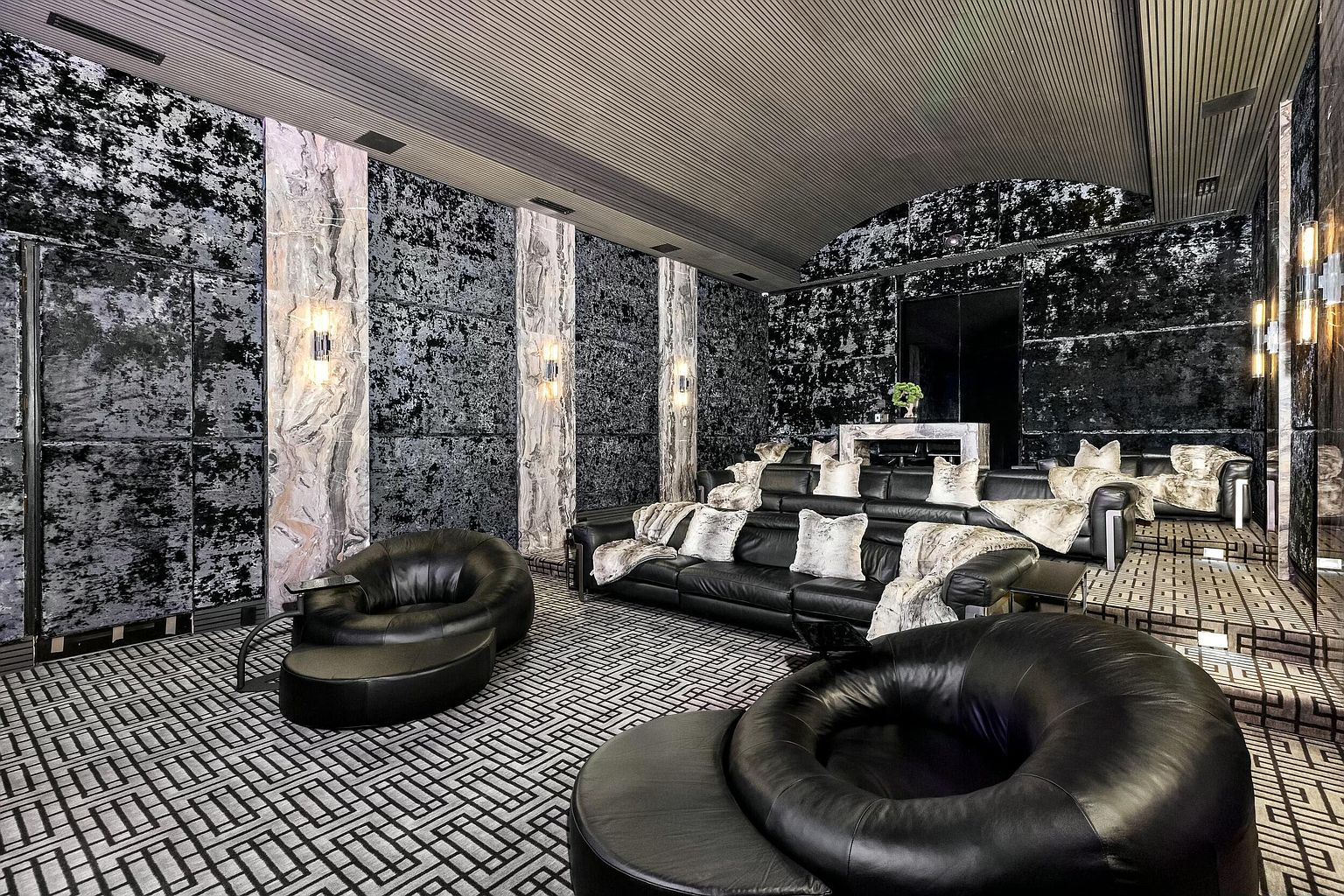 This is a luxurious living room featuring black leather seating, including a sectional sofa adorned with plush pillows and throws, and unique round chairs. The walls are decorated with a textured black material and marble-like columns with sconces, creating a sophisticated and dramatic ambiance. The geometric patterned carpet adds a modern touch to the space.