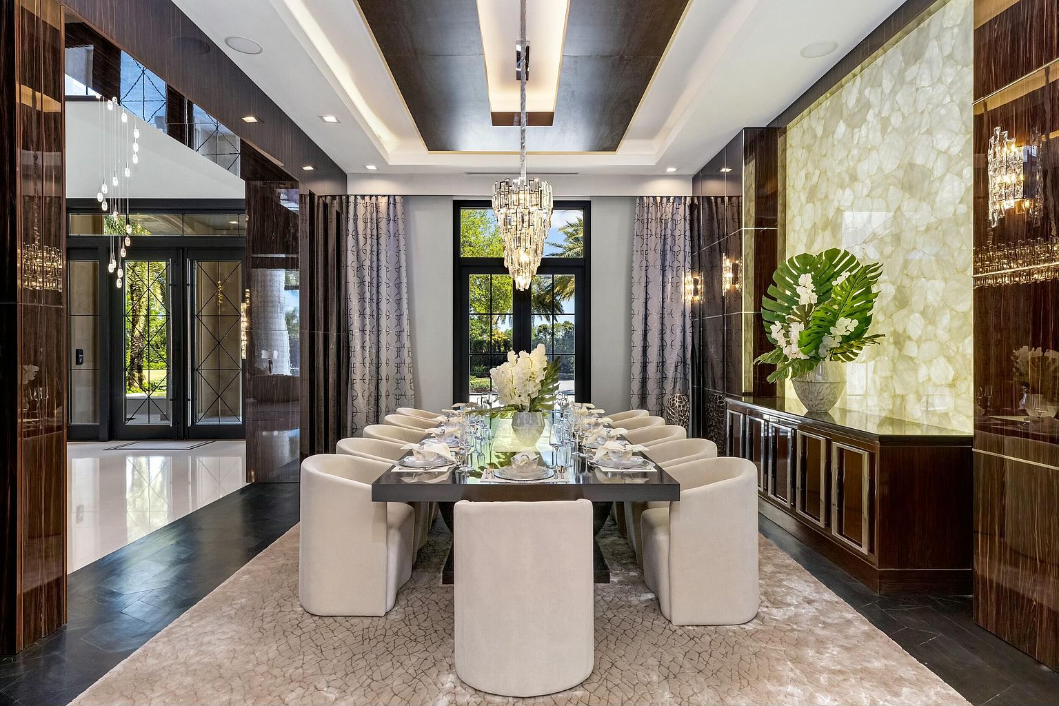 This is a luxurious dining room featuring a long, rectangular dining table with seating for twelve, surrounded by elegant, upholstered chairs. A modern chandelier hangs above the table, and the room is decorated with sophisticated curtains and a decorative wall feature. The overall impression is one of opulence and refined taste, perfect for formal gatherings.