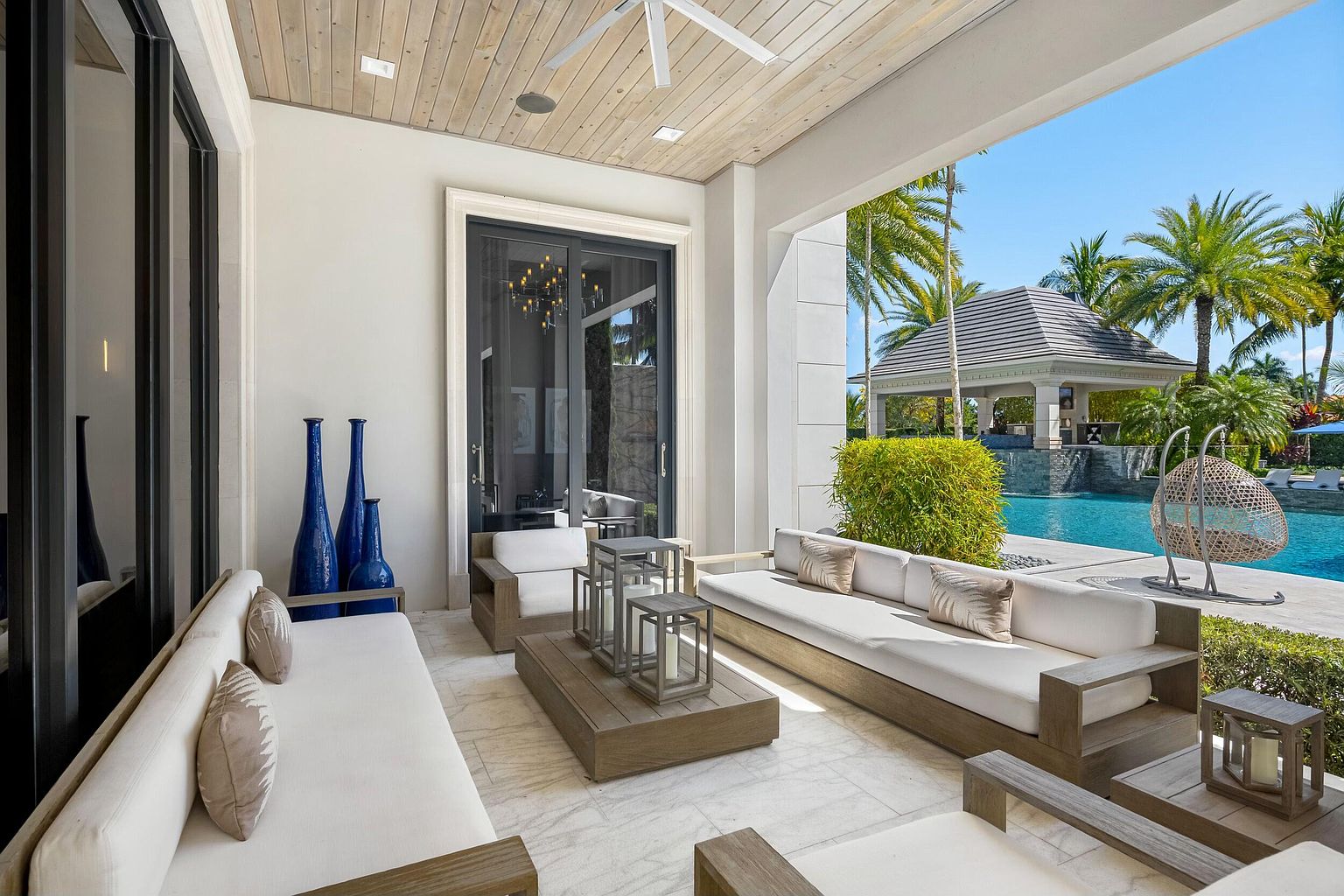 This image showcases a luxurious outdoor patio area, featuring comfortable seating arrangements with plush cushions and stylish wooden frames. The patio overlooks a stunning pool and a gazebo, surrounded by lush tropical landscaping. The space is designed for relaxation and entertainment, offering a seamless transition between indoor and outdoor living.