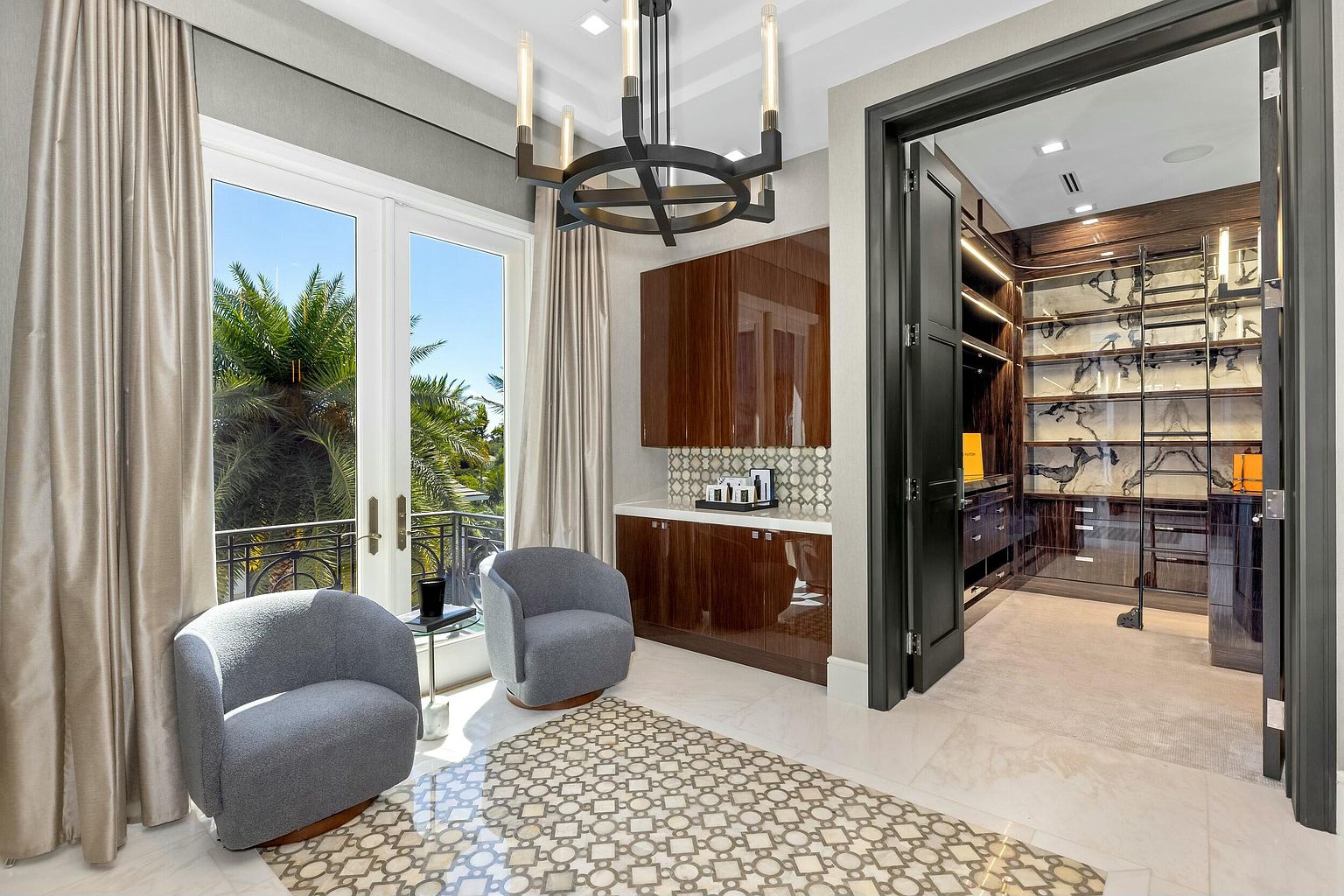 This image showcases a luxurious walk-in closet adjacent to a seating area with two armchairs and a balcony. The closet features custom shelving, glass displays, and elegant lighting, creating a high-end boutique feel. The seating area offers a comfortable space to relax and enjoy the view, enhancing the overall appeal of the room.