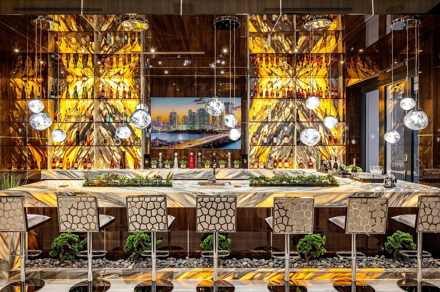This image showcases a luxurious dining area featuring a well-stocked bar with illuminated shelves displaying various bottles. The bar is complemented by stylish bar stools with unique patterned backs, and the space is adorned with modern, spherical pendant lights. The overall impression is one of sophistication and high-end design, perfect for entertaining.