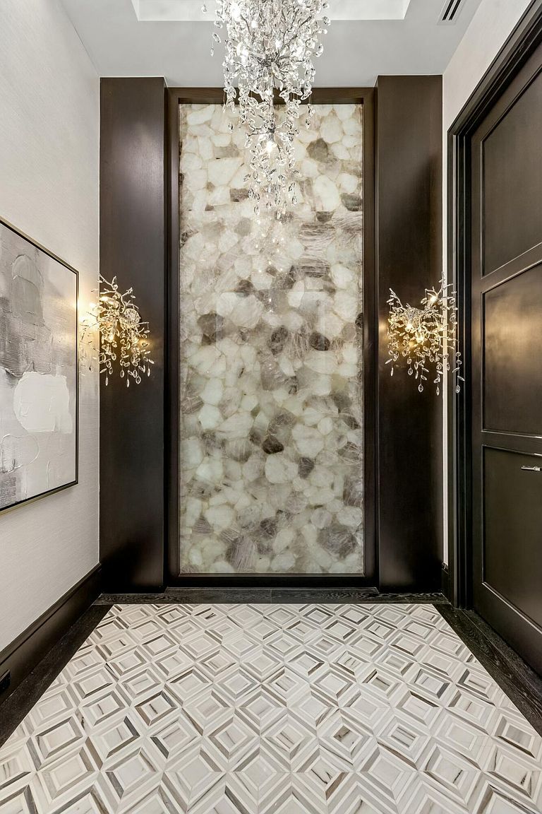 This interior shot showcases a luxurious hallway featuring a striking accent wall made of what appears to be backlit agate or a similar stone material, flanked by dark wood panels and sconces. The floor is tiled in a geometric pattern, and a crystal chandelier hangs from the ceiling, adding to the opulent feel. A dark wood door is visible on the right, suggesting a transition to another room.