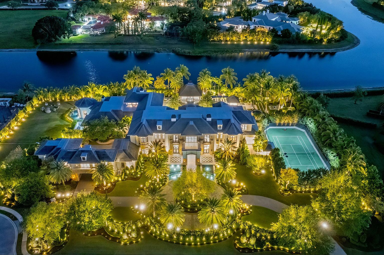 This aerial view showcases a luxurious estate at night, featuring a grand mansion with a symmetrical design, a tennis court, and meticulously landscaped grounds illuminated by numerous lights. The property is bordered by a body of water, adding to its exclusivity and appeal. The cinematic perspective highlights the scale and opulence of the property.