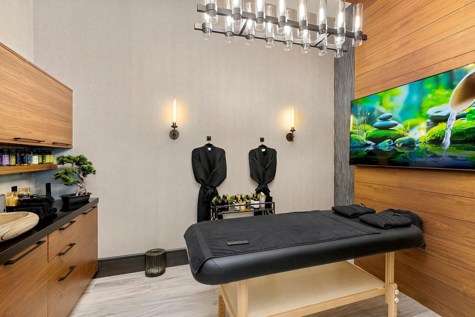 This interior shot showcases a serene spa or massage room, designed for relaxation and wellness. The room features a massage table with black linens, flanked by two black robes hanging on the wall, and a modern light fixture overhead. A flat-screen TV displays a calming nature scene, complemented by wood paneling and soft lighting, creating a tranquil and inviting atmosphere.