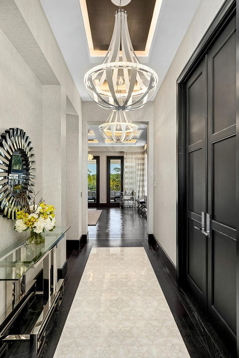 This interior shot showcases a luxurious hallway with dark hardwood floors and a patterned runner. A modern chandelier hangs from the ceiling, and a mirrored console table with floral arrangement adds elegance. The hallway leads to a bright living space, creating a sense of depth and sophistication.
