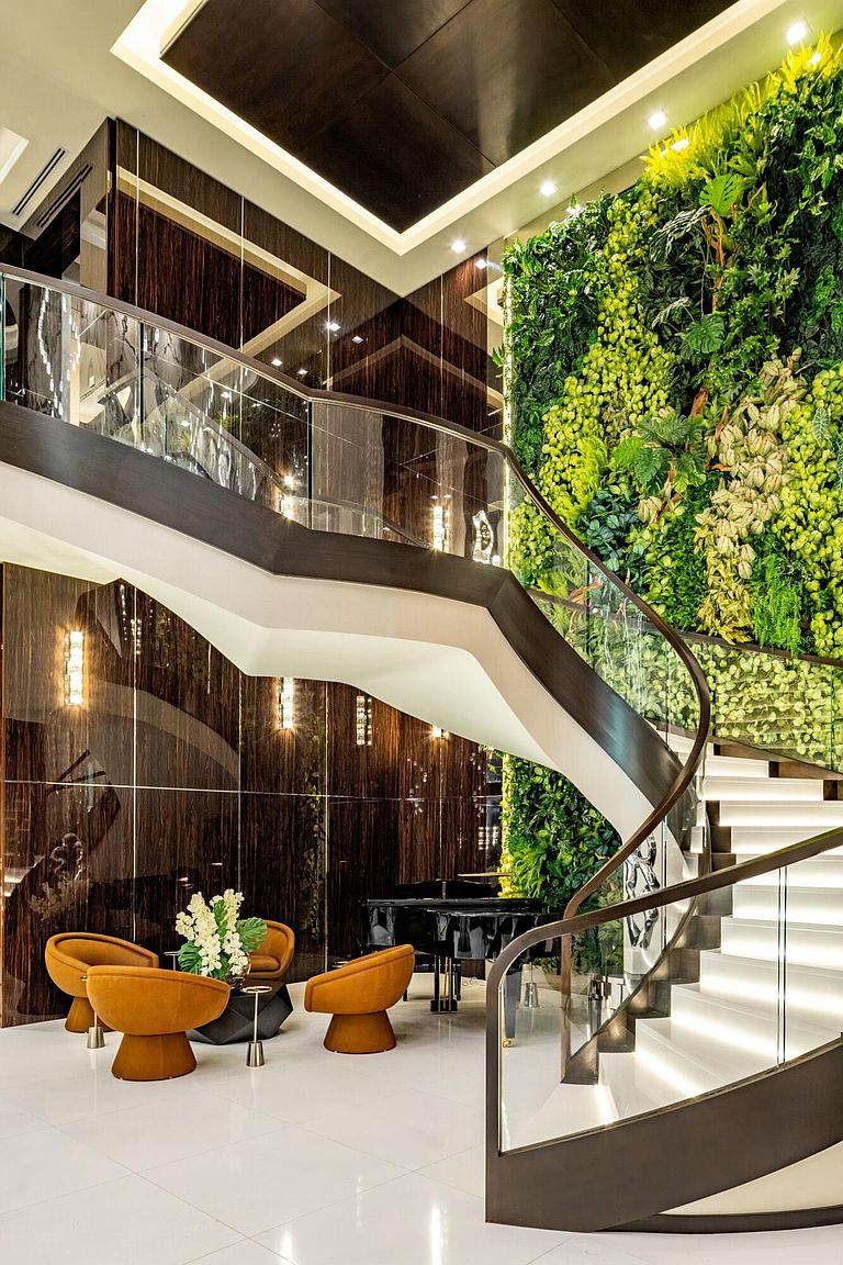 This is a luxurious interior shot featuring a grand, curved staircase with glass railings and illuminated steps. A striking green wall adds a natural element, while elegant seating and a piano create a sophisticated lounge area. The overall impression is one of opulence and modern design.