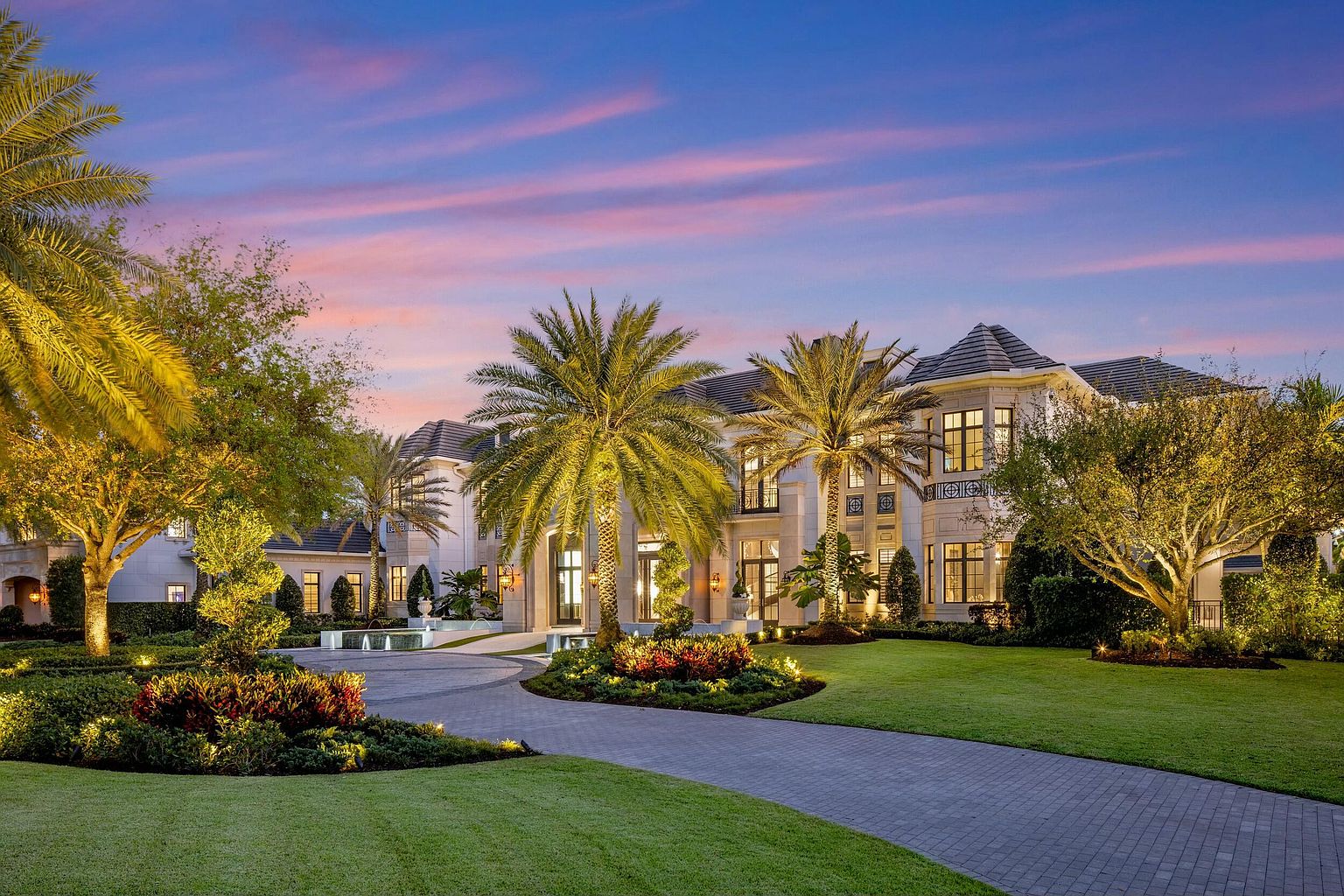 This is a stunning front view of a luxurious estate at dusk, showcasing its grand architecture and meticulously landscaped grounds. The property features a circular driveway, lush green lawns, mature palm trees, and vibrant flower beds, all illuminated by strategically placed lighting. The overall impression is one of elegance, opulence, and curb appeal.