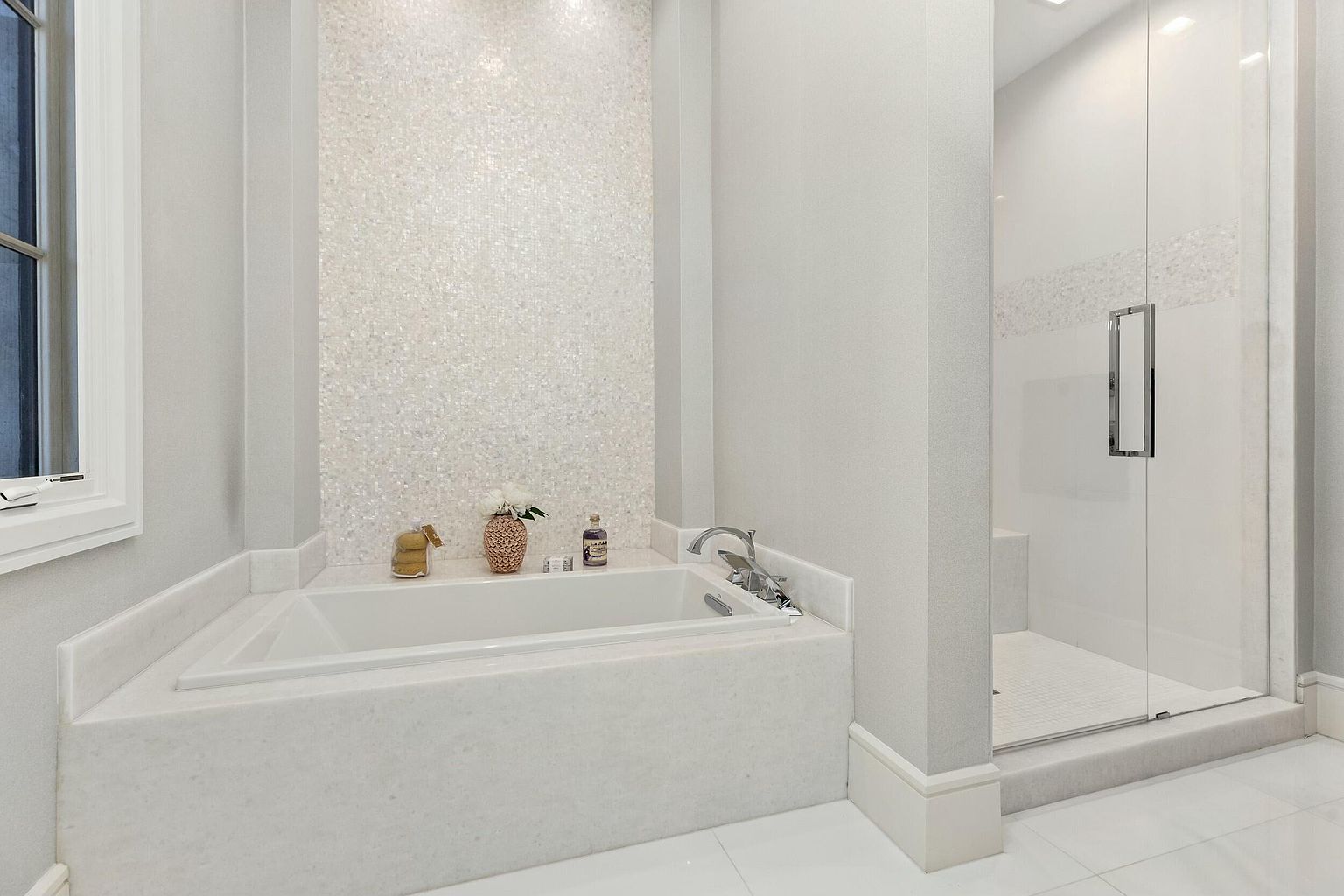 This is a luxurious primary bathroom featuring a soaking tub with a white marble surround and a textured, shimmering accent wall. A glass-enclosed shower is visible to the right, contributing to the spa-like atmosphere. The color palette is predominantly white, creating a clean and serene space.