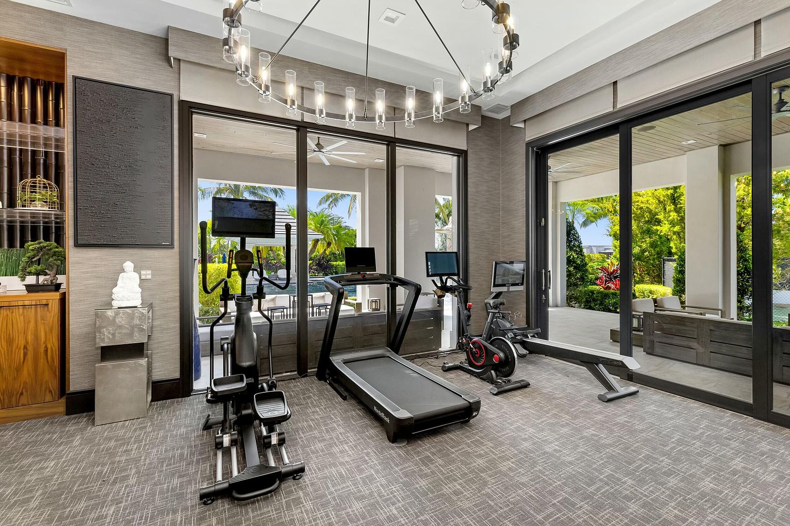 This is an interior shot of a modern home gym. The room features an elliptical machine, treadmill, stationary bike, and rowing machine, all arranged on a gray patterned carpet. Large sliding glass doors offer a view of the outdoor patio and garden, while a contemporary chandelier hangs from the ceiling, creating a bright and inviting workout space.