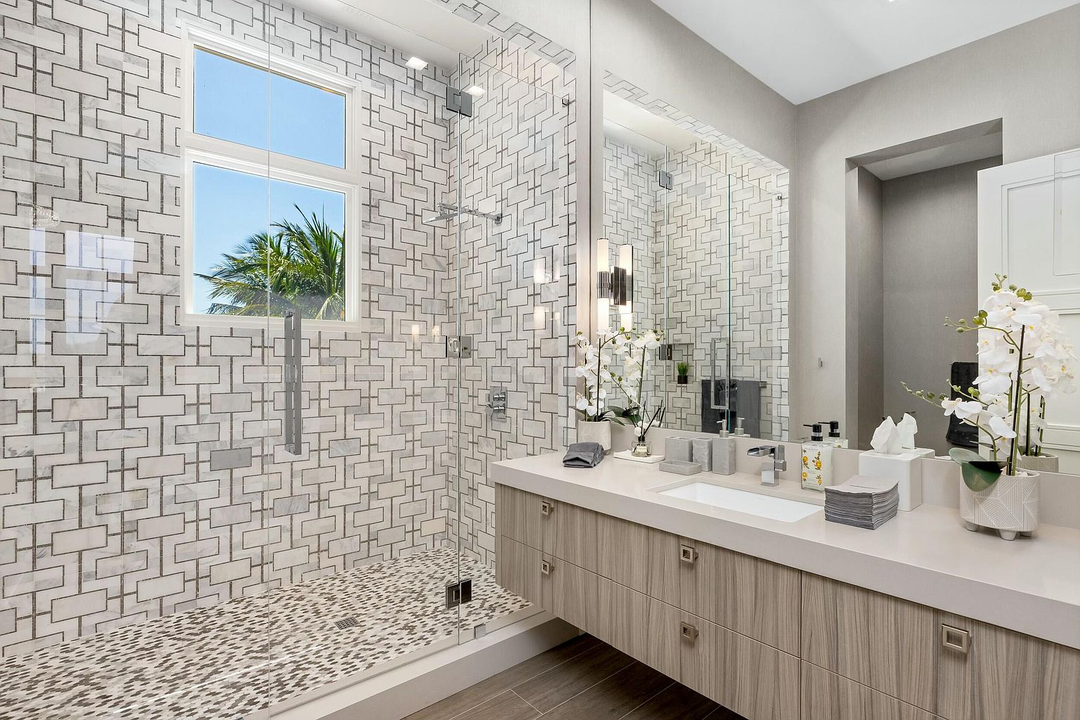 This is a luxurious primary bathroom featuring a glass-enclosed shower with patterned tile walls and a pebble floor. A large vanity with a light-colored countertop and wood-grain cabinets provides ample storage and counter space. The room is well-lit and exudes a spa-like atmosphere, perfect for relaxation.