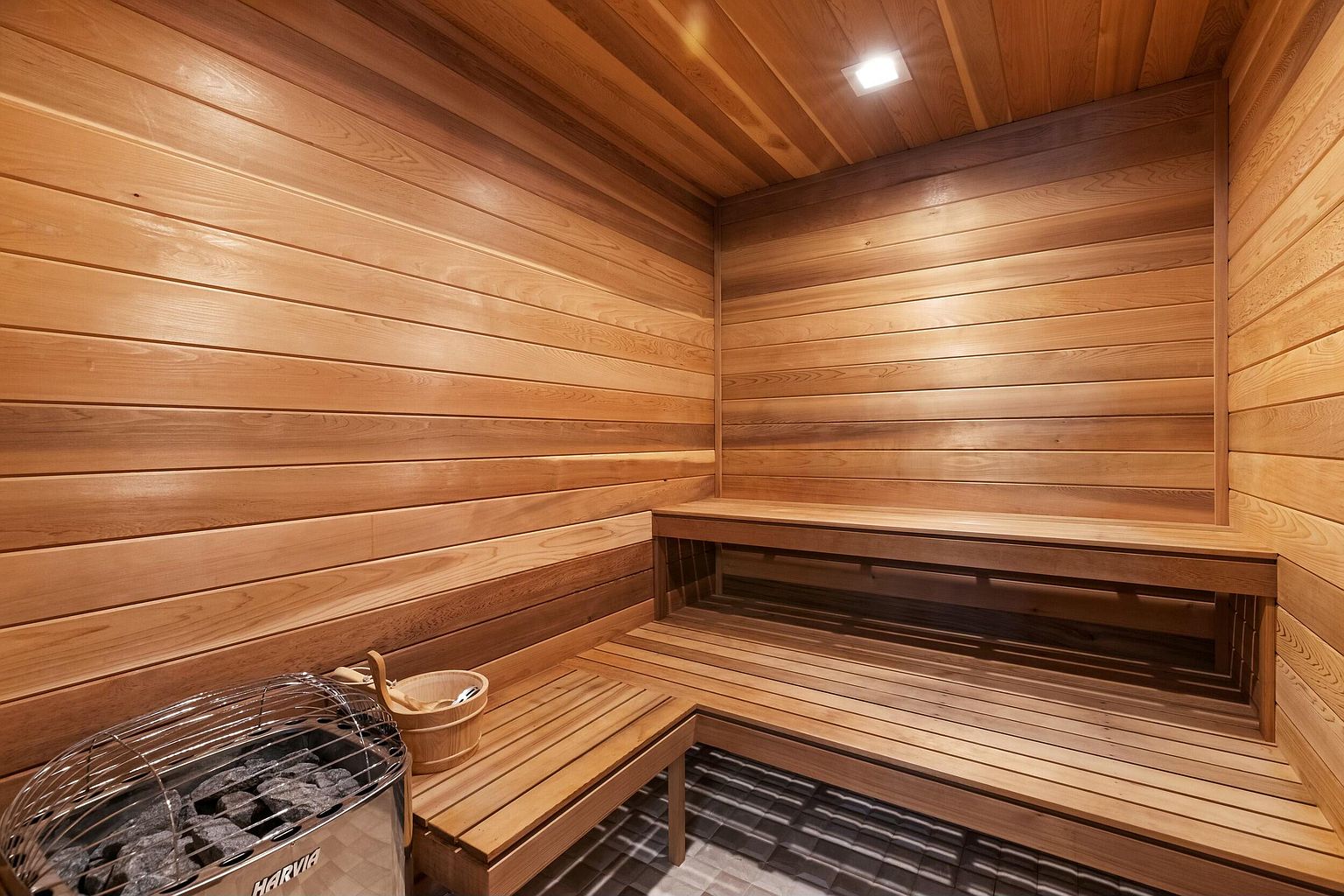 This is an interior shot of a sauna, featuring wooden walls, ceiling, and benches. A metal sauna heater with rocks sits in the corner, along with a wooden bucket and ladle. The room is well-lit, creating a warm and inviting atmosphere, perfect for relaxation and wellness.