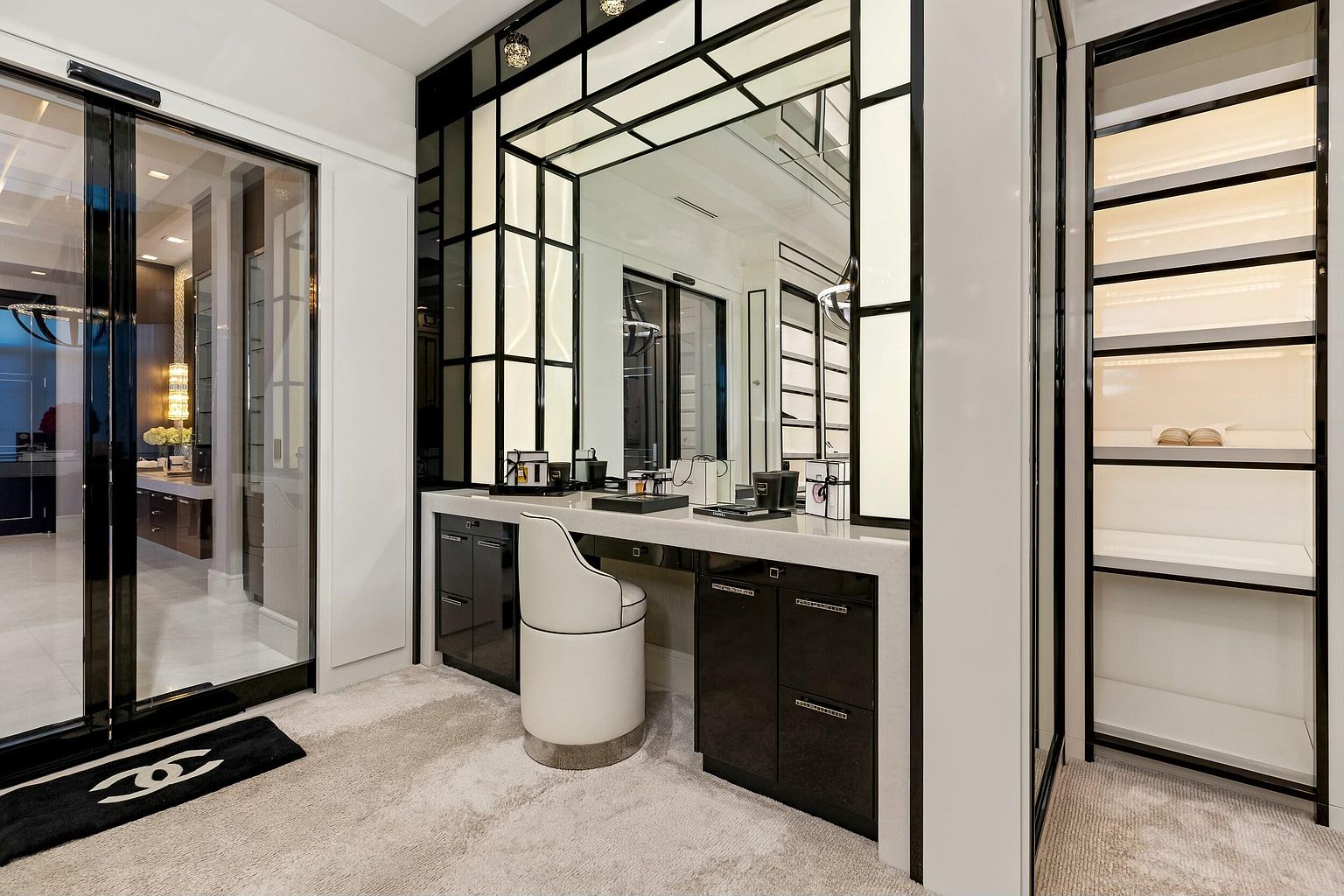 This is a luxurious walk-in closet featuring custom cabinetry in a black and white color scheme. A vanity area with a large mirror and a stylish chair is the focal point, complemented by ample storage space with illuminated shelves. The overall impression is one of high-end design and organization.