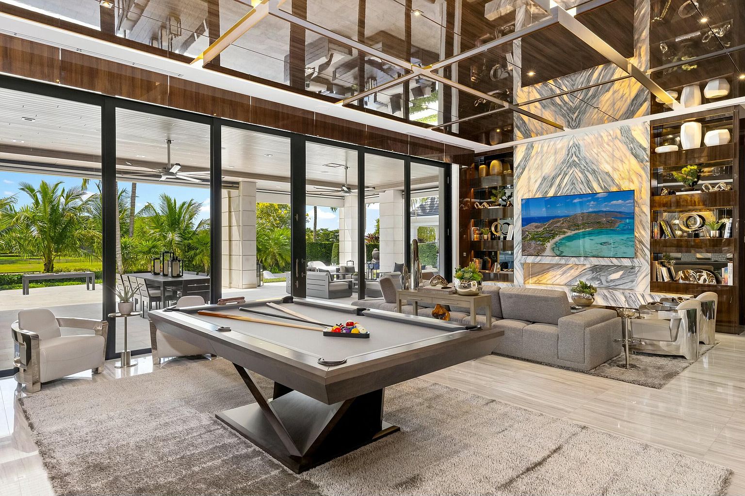 This is a luxurious living room featuring a pool table as a central element, complemented by a modern gray sofa and stylish armchairs. The room boasts a high, reflective ceiling and a large marble-clad wall with a built-in entertainment center. Large sliding glass doors offer a view of the outdoor patio and lush landscaping, creating a seamless indoor-outdoor living experience.