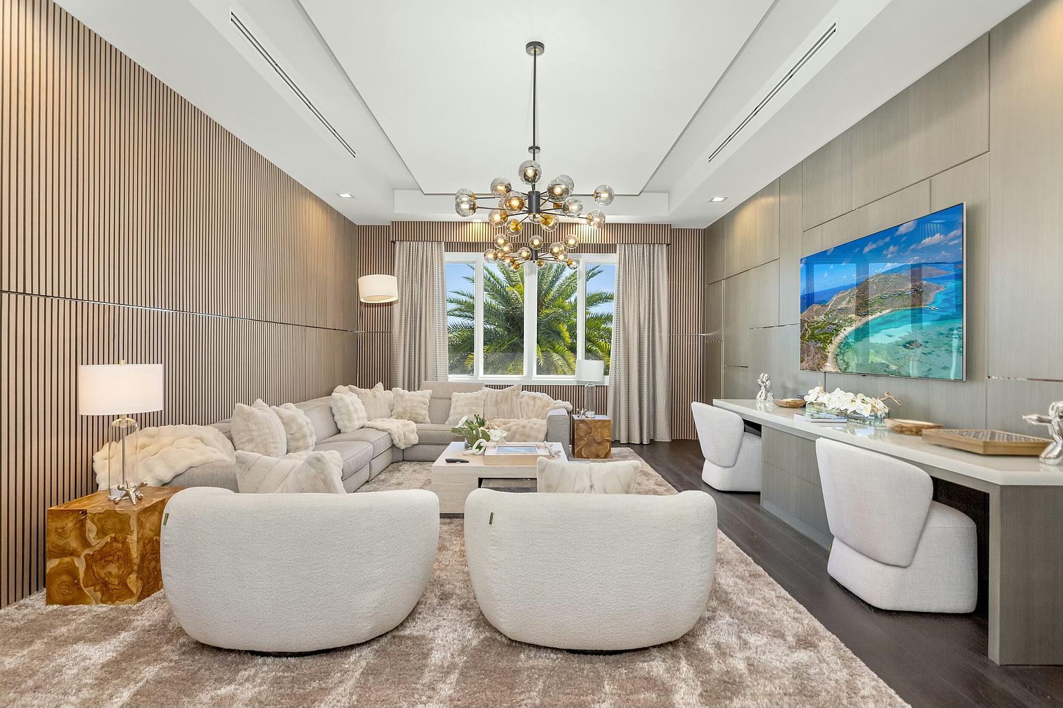 This is a luxurious living room featuring a large sectional sofa adorned with plush pillows, two rounded white armchairs, and a modern chandelier. The room is decorated with wood paneling and a large television, creating a sophisticated and comfortable atmosphere. The space is well-lit and appears spacious, perfect for relaxation and entertainment.