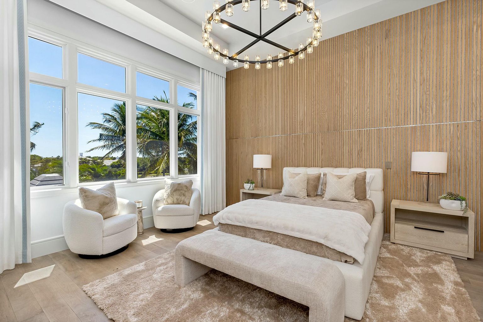 This is a luxurious primary bedroom featuring a king-size bed with neutral-toned bedding, flanked by matching nightstands and lamps. A striking circular chandelier hangs above, complementing the wood-paneled accent wall. Two cozy armchairs sit near a large window offering a view of palm trees, creating a serene and inviting atmosphere.