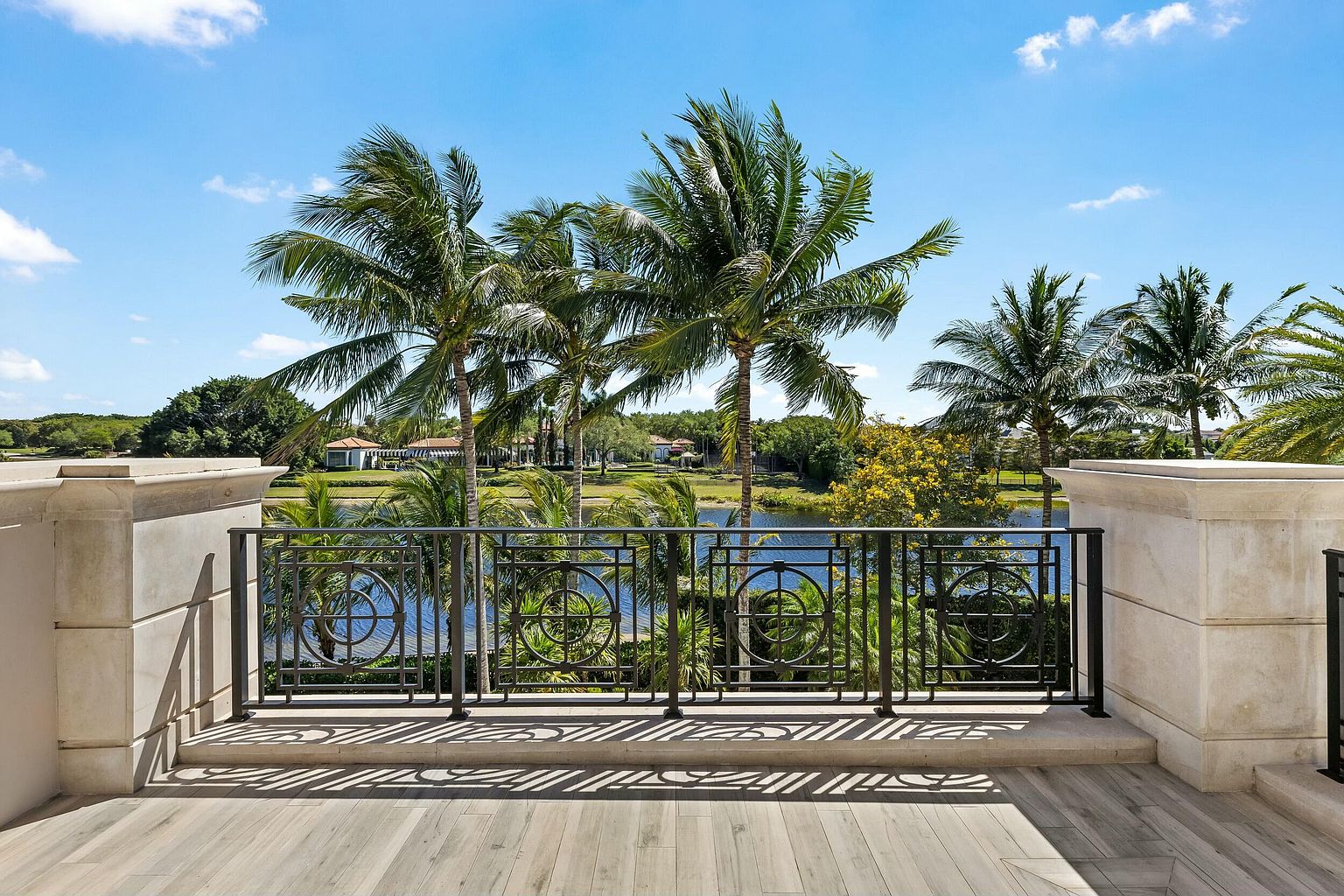 This image showcases a luxurious balcony overlooking a serene lake and lush palm trees. The balcony features a decorative wrought iron railing and elegant stone pillars, complemented by a stylish wooden deck. The view offers a tranquil and upscale ambiance, perfect for relaxation and enjoying the natural surroundings.