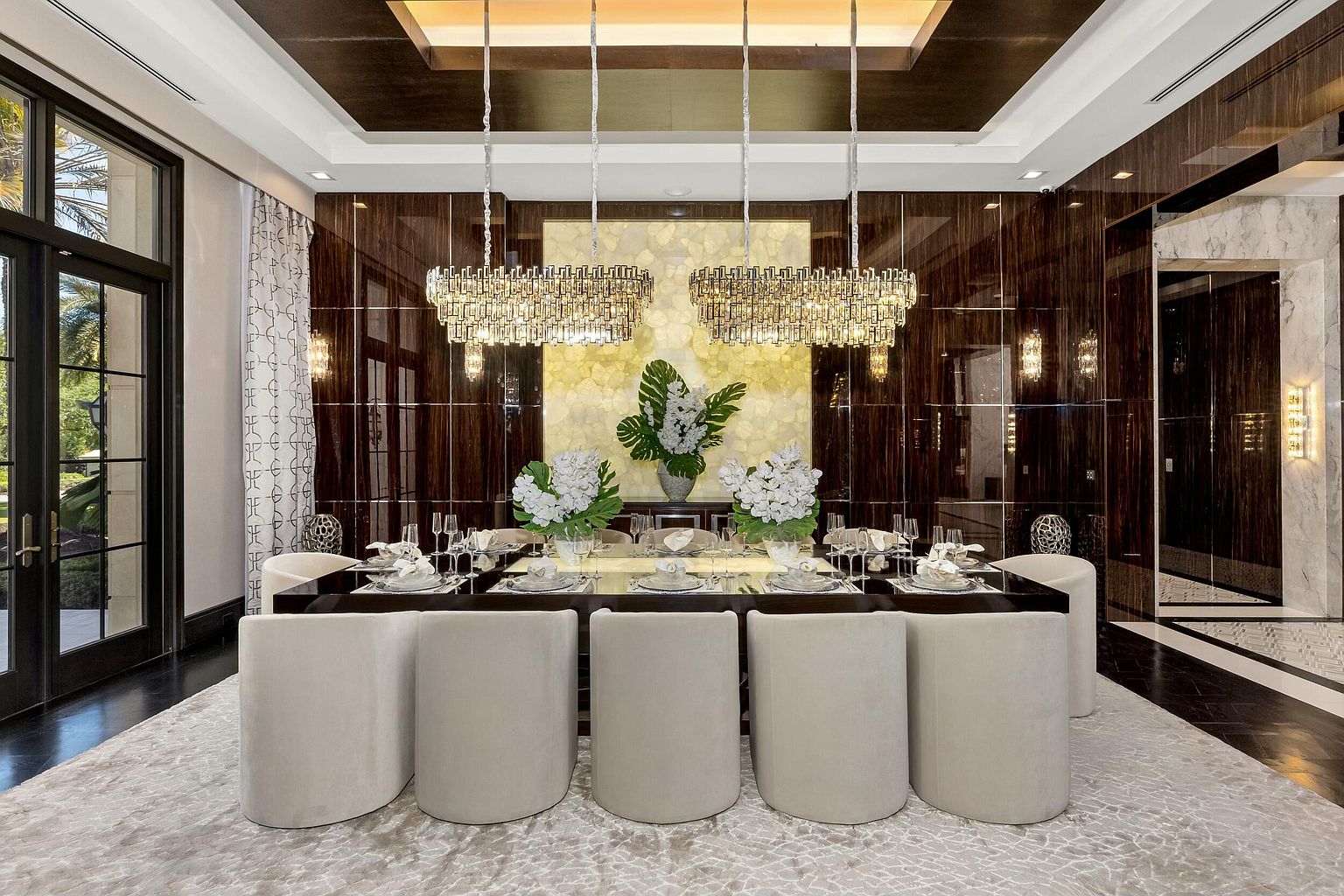 This is a luxurious dining room featuring a long, dark wood table set for a formal meal, surrounded by elegant, curved beige chairs. Two large, ornate chandeliers hang above the table, illuminating the space and highlighting the glossy wood paneling on the walls. The room is grounded by a large, patterned area rug, and the overall impression is one of sophistication and opulence.