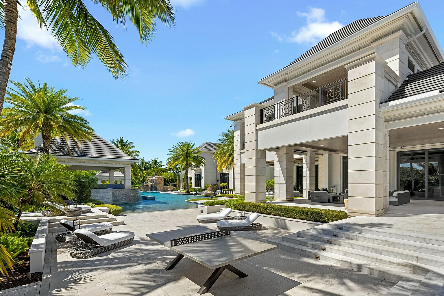 This image showcases a luxurious backyard featuring a large pool with a waterfall feature, surrounded by lush tropical landscaping and palm trees. A covered pavilion sits adjacent to the pool, and a modern, multi-level patio area includes lounge chairs and a ping pong table. The architectural style of the main house is grand and contemporary, with stone cladding and a balcony overlooking the pool area.