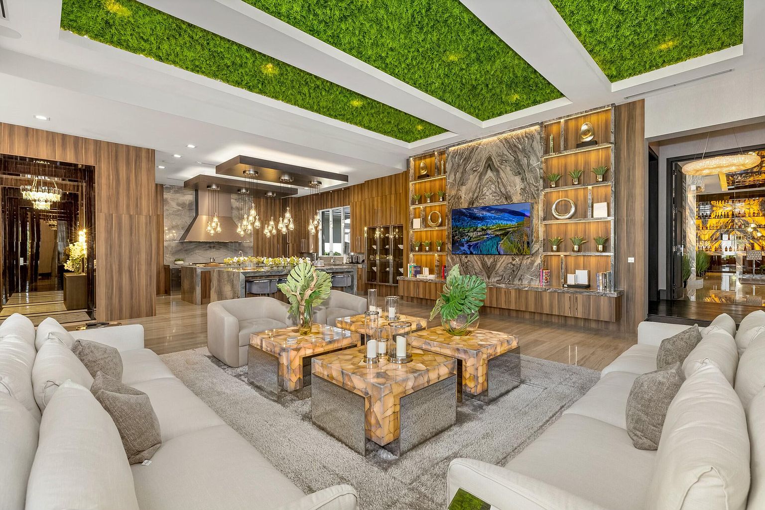This is a luxurious living room featuring a unique moss-covered ceiling with recessed lighting, creating a natural and modern ambiance. The room is furnished with plush white sofas, geometrically designed coffee tables, and a large decorative wall with built-in shelving and a flat-screen TV. The open-concept design seamlessly connects the living area to the kitchen, enhancing the sense of spaciousness and upscale living.